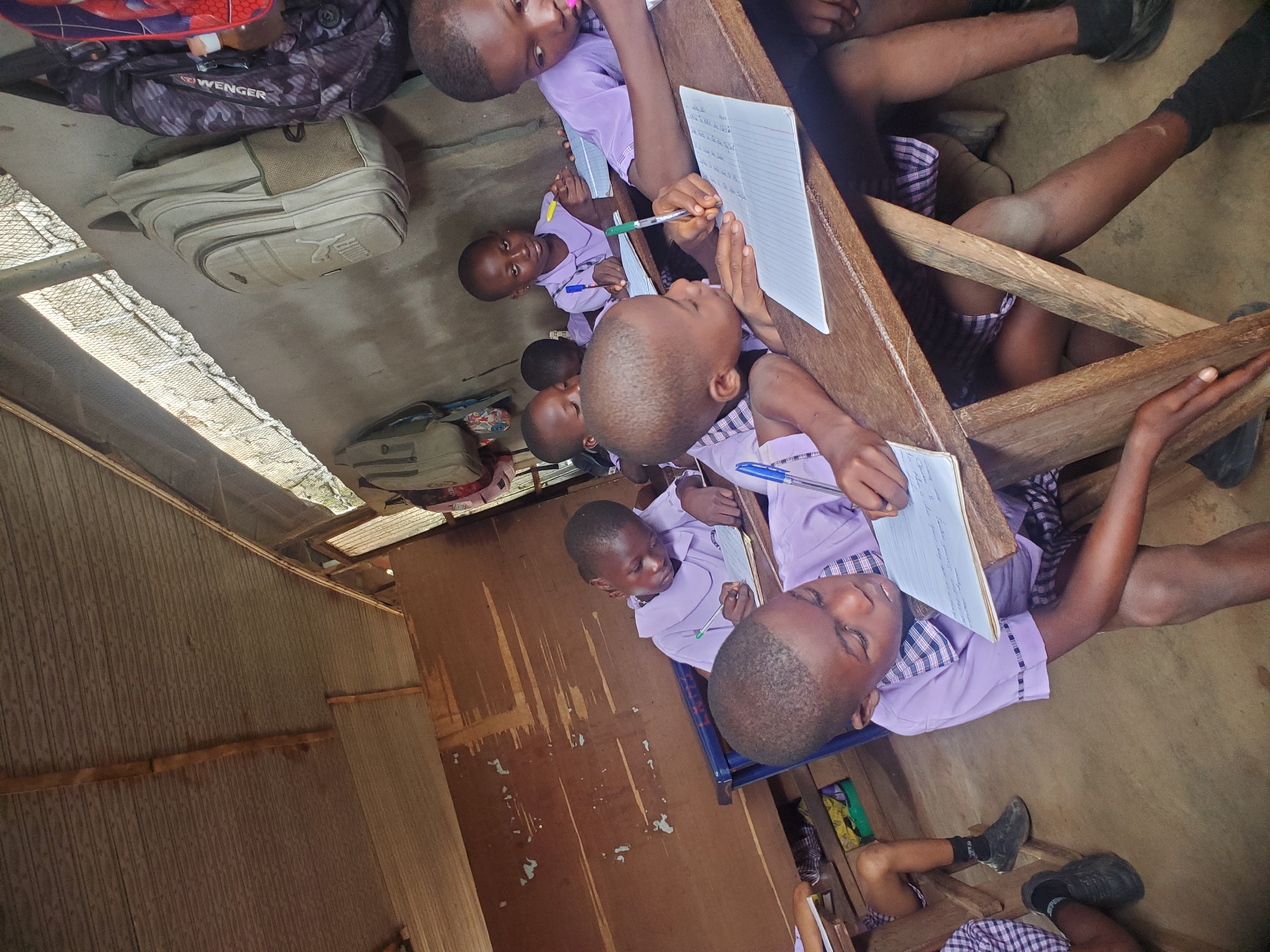 In a classroom in Ukwu-Abwa, Nigeria, several young students in lavender and checkered uniforms are seated at wooden desks, writing in notebooks with pens. Backpacks are strewn on the floor near the desks. The room has basic construction, with simple walls and a visible corrugated metal roof. The students appear engaged in their work.