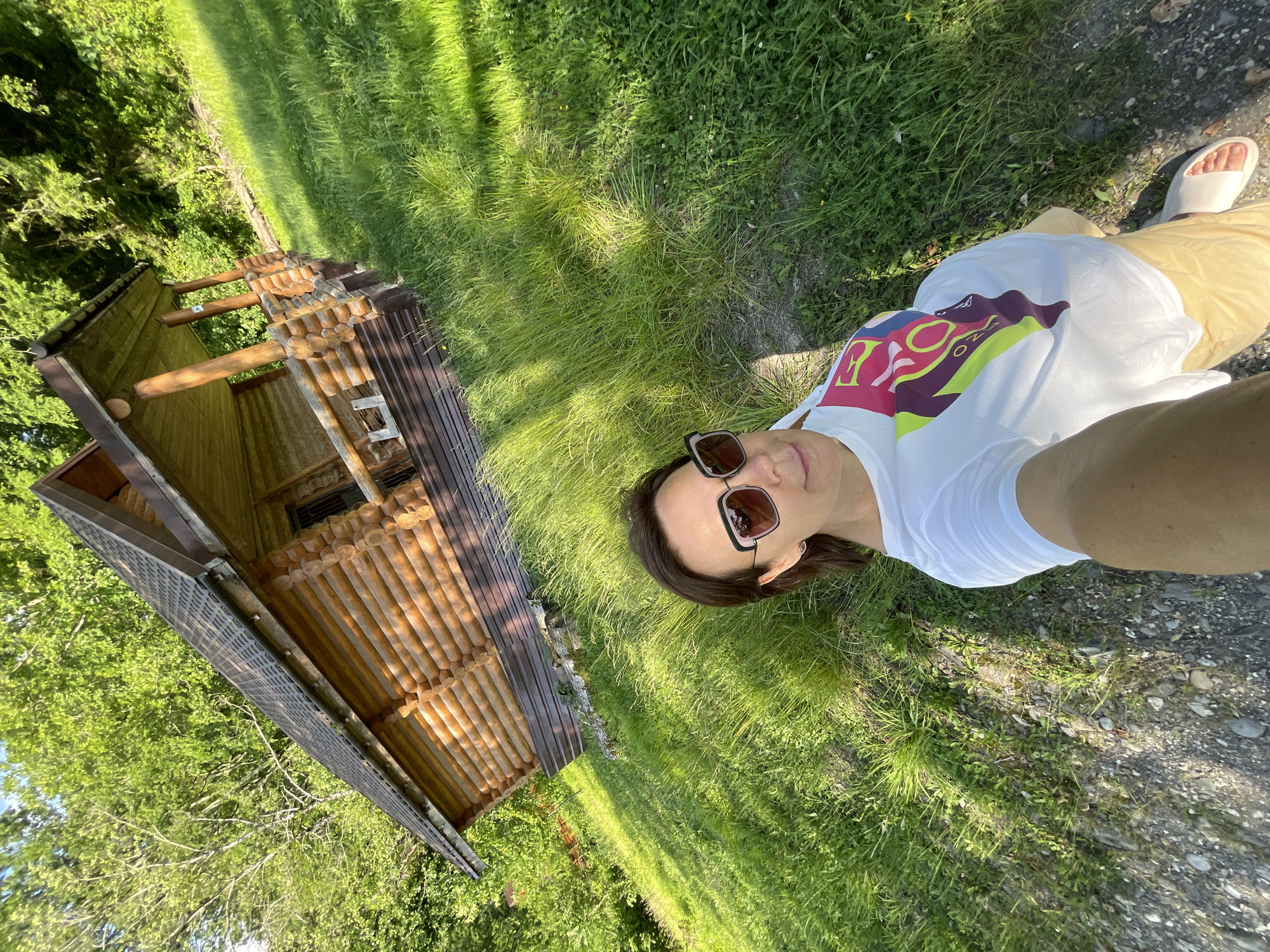 An adult female is depicted taking a selfie outdoors on a sunny day in the city of Sochi, Russia. She has short, dark hair and is wearing dark-framed sunglasses with light-colored lenses. Her attire consists of a white short-sleeved t-shirt with a colorful, abstract graphic design on the front, light-colored (likely beige or yellow) pants, and white slide sandals. She holds the camera at arm's length, angling it to include herself, a grassy slope, and a log cabin in the background.
The log cabin, constructed from light-brown, horizontally stacked wooden logs, features a steeply pitched dark roof with a visible gutter system. A small, covered porch supported by several rounded wooden posts extends from the front of the cabin. The lower section of the cabin's exterior is covered in a dark brown material. The cabin is situated on a lush green hillside, with the grass appearing somewhat overgrown in areas. Dense green trees form a forest behind the cabin and further up the slope. In the immediate foreground, near the person, the ground is a mix of dirt, small pebbles, and sparse green vegetation. Bright sunlight casts distinct shadows, indicating clear weather.