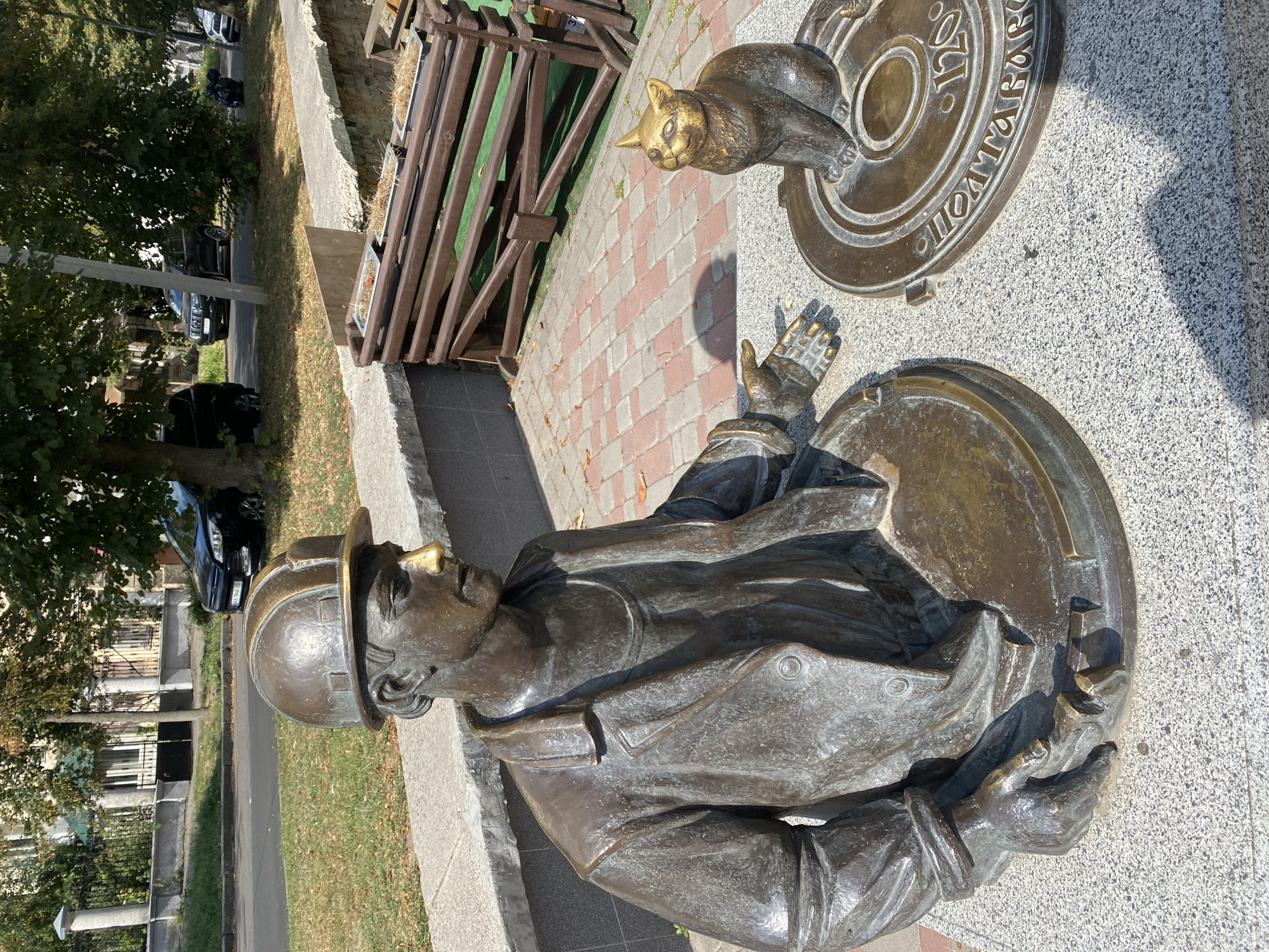 This is a photograph of a bronze sculpture of a construction worker and a cat, located in Poltava, Ukraine. The sculpture depicts the worker, wearing a hard hat and a coat, emerging from a manhole. He is pointing towards a golden-colored cat sitting on another manhole cover. The cat is looking up at the worker with its mouth slightly open. The manhole covers have inscriptions on them. The sculpture is situated outdoors, on a paved area with a brick pattern, next to a low stone wall. In the background, there are trees, a street with cars, and a building. The weather appears to be sunny.