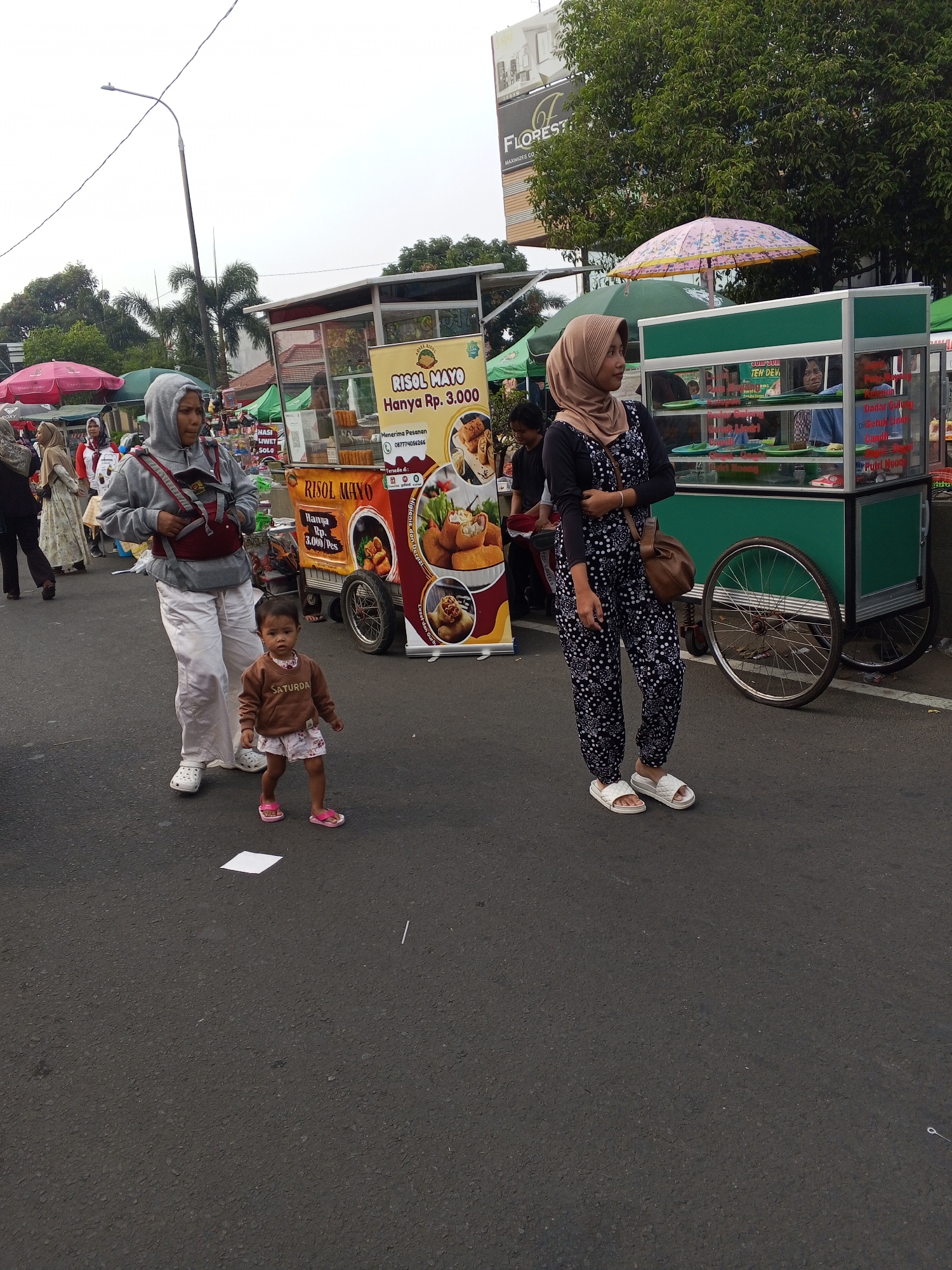 In the city of Serang, Indonesia, on a paved street, two women and a young child are captured in a full shot. The woman on the left, wearing a gray hoodie and white pants, is seen from the waist up, carrying a baby in a red carrier. She is looking down at the child, who is facing the camera. The child, dressed in a brown sweater with "Saturday" printed on it and pink shorts, stands in front of her, looking forward.
To the right, another woman, dressed in a beige hijab and a black and white polka-dot jumpsuit, stands with her left hand on her chest, looking to the left. She wears white slides on her feet and a brown crossbody bag.
Behind them, a food cart is visible, displaying "RISOL MAYO" on its signage. The cart is green and white, with a large wheel on the right side. Another cart with a pink umbrella is partially visible to the left. In the background, trees and a billboard with the word "FLOREST" are seen. The overall scene suggests a lively street market or food stall area.