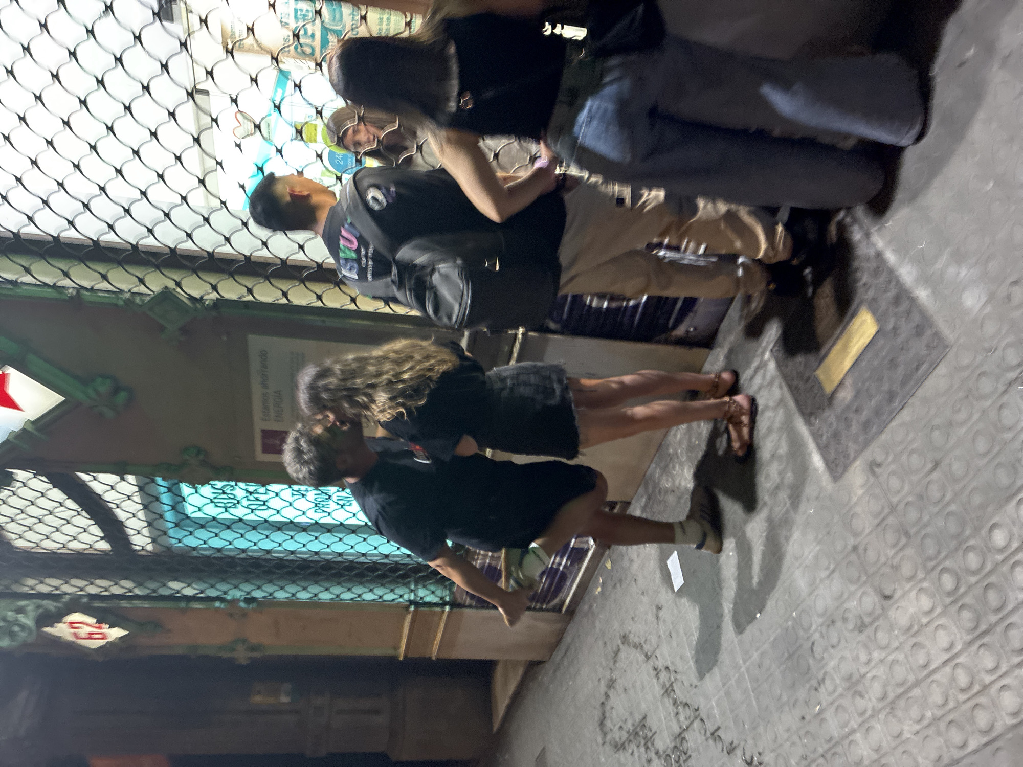 The image depicts a nighttime street scene in Barcelona, Spain, identifiable by the distinctive "panot" (flower tile) sidewalk pattern and modernist architectural elements. The scene is illuminated by artificial light, casting soft shadows.
In the center, a couple is kissing in front of a building with a ornate green facade and a security grille covering a shop window. The male, positioned on the left, wears a black t-shirt, black shorts, white socks, and light-colored sneakers; he is leaning forward, with his left leg bent and raised behind him, indicating a moment of romantic embrace. The female, on the right, wears a black t-shirt, a dark mini-skirt, and light-brown sandals with ankle straps; she has long, wavy brown hair.
To the right of the kissing couple, three other individuals are standing near another shop window, also covered by a security grille. The first person in this group is a male with short, dark hair, seen from behind, wearing a dark t-shirt with colorful lettering, light-colored pants, and a black backpack. To his right, a female with medium-length dark hair is partially visible, looking towards the shop interior. The furthest individual on the right is a female, seen from behind, wearing a black tank top and blue jeans, with a black bag slung across her body.
The building facade features decorative green elements and a red and white diamond-shaped sign with the number "62." A partially visible sign behind the kissing couple reads "Estamos ahorrando ENERGIA" (We are saving energy), confirming the Spanish language. Another sign visible through the grille behind the right group indicates "24h" and "OPEN." The street surface is a patterned tile sidewalk, appearing somewhat damp in areas, with a discarded white paper visible near the center.
