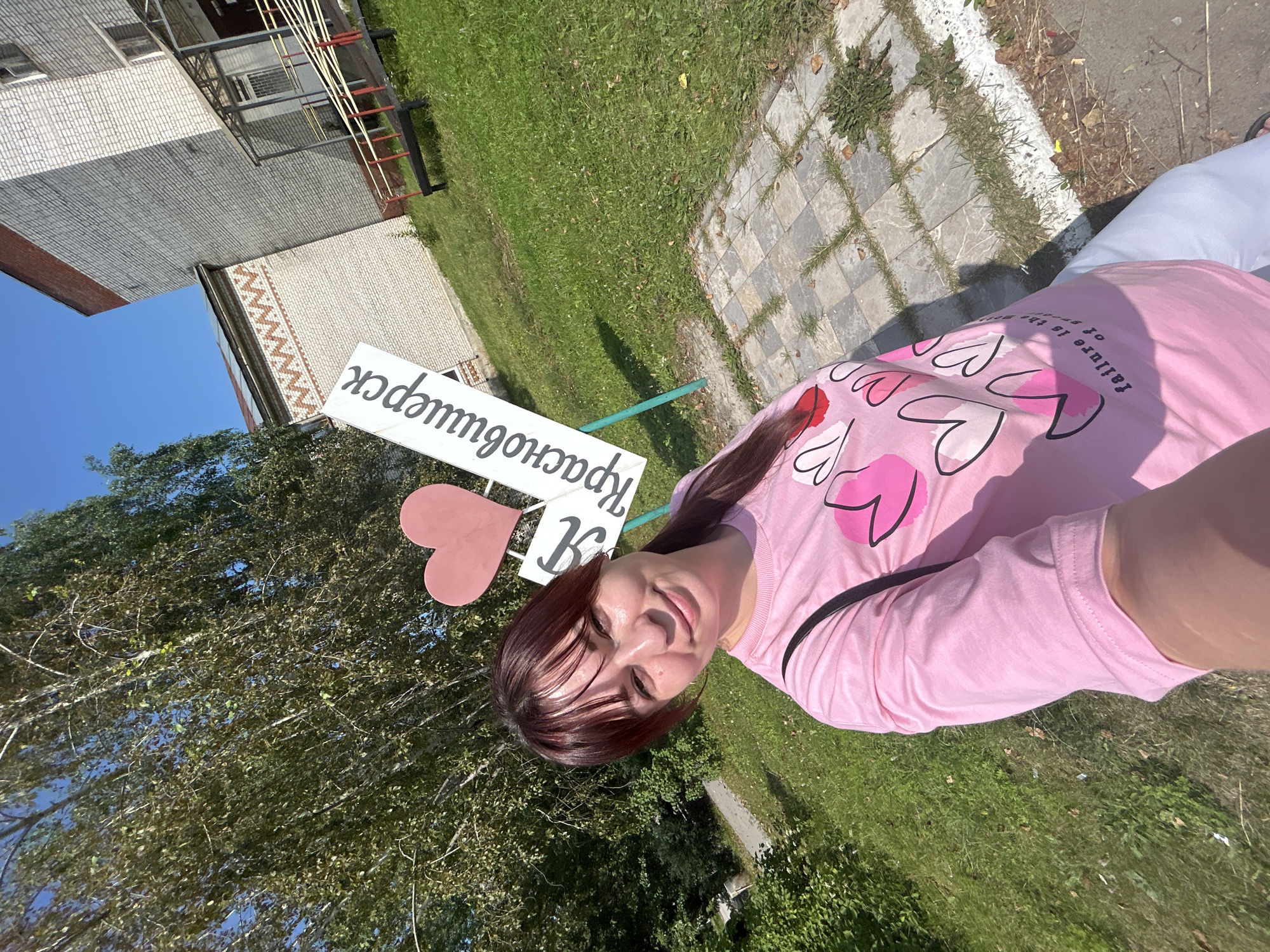 A young woman with dark hair and bangs, wearing a pink t-shirt with hearts on it, smiles at the camera. She is standing outdoors in what appears to be a park or public space. Behind her, a white sign with a pink heart and Cyrillic text reads "Я люблю Красновишерск" (I love Krasnovishersk), indicating the location is the city of Krasnovishersk, Russia. The background includes green grass, trees, and a brick building with an awning. The woman is taking a selfie.
