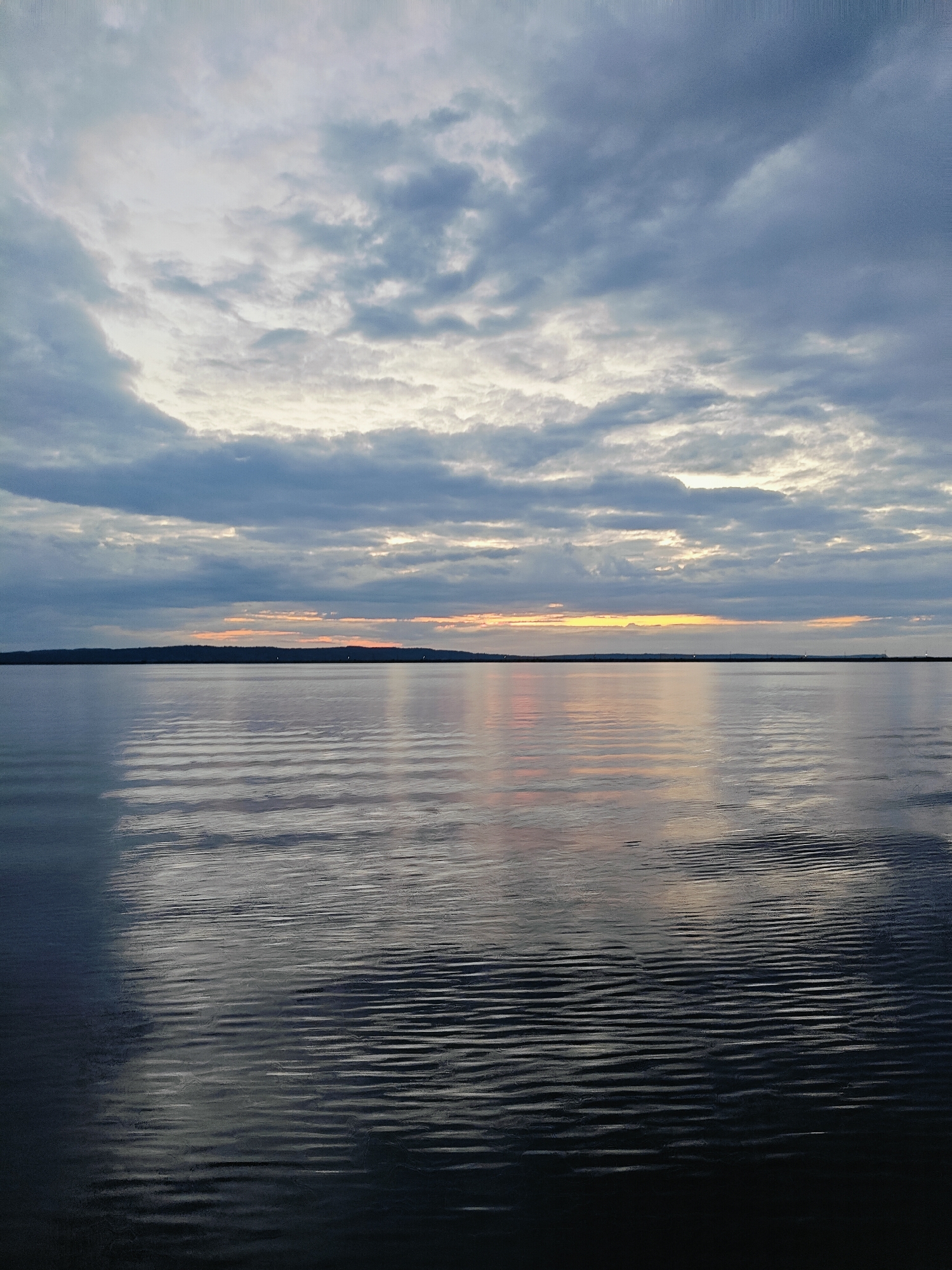 This is a wide-angle, full-frame photograph of a body of water, likely the Kama River in Chaikovsky, Russia, under a cloudy sky. The water's surface exhibits a ripple pattern, with the reflections of the sky and a distant dark treeline visible. The sky is filled with layered clouds, ranging in color from deep gray to lighter shades, with a band of soft orange and yellow light near the horizon, suggesting either sunrise or sunset. The overall mood is serene and expansive. There are no people, animals, or man-made structures clearly visible in the image. The composition is balanced, with the horizon line dividing the sky and water approximately in half.