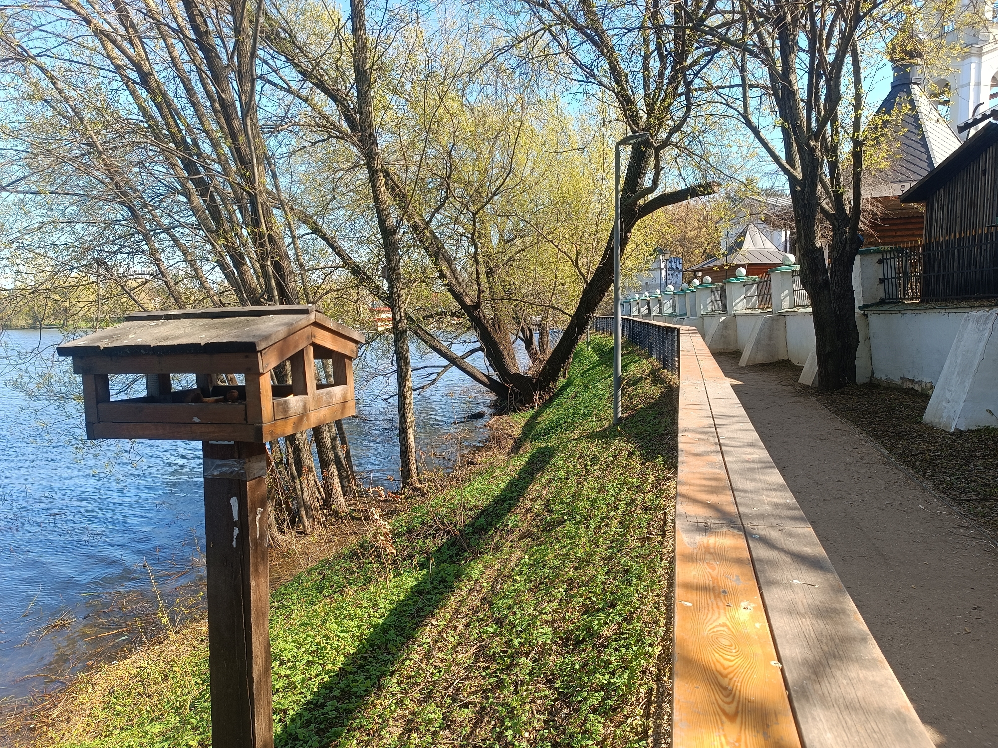 The image shows a view of the Krutitsy Patriarchal Metochion in Moscow, Russia. A wooden bird feeder stands in the foreground on the left side of the frame. Behind it is a body of water, likely the Moskva River or a connected pond. On the right, a wooden walkway runs parallel to a white wall and fence. Beyond the fence, parts of the Metochion buildings, including a church with a steeple, are visible. The trees are in early spring foliage, with some buds and new leaves appearing. The ground is covered in green vegetation and shadows cast by the trees and structures. No people are visible in the image.