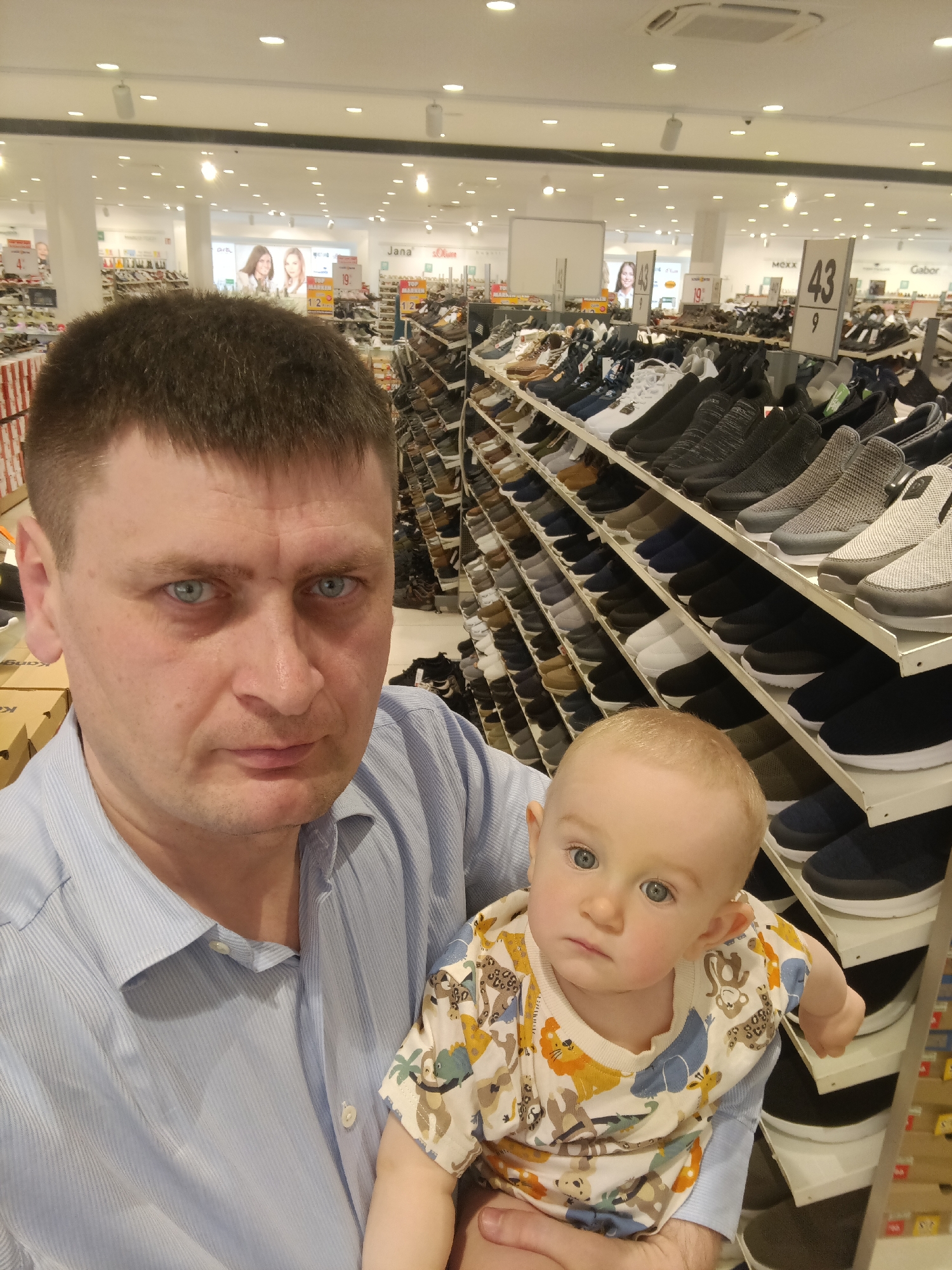 In a shoe store in Nuremberg, Germany, a man with short brown hair and blue eyes, wearing a light blue striped button-down shirt, holds a baby wearing a light-colored t-shirt with an animal print. The man's face is close to the camera, while the baby is positioned slightly further away, looking towards the camera. In the background, rows of shelves are filled with various shoes. The store appears well-lit with overhead fluorescent lighting. There are signs visible in the background with numbers and brand names, indicating this is a retail environment for footwear.