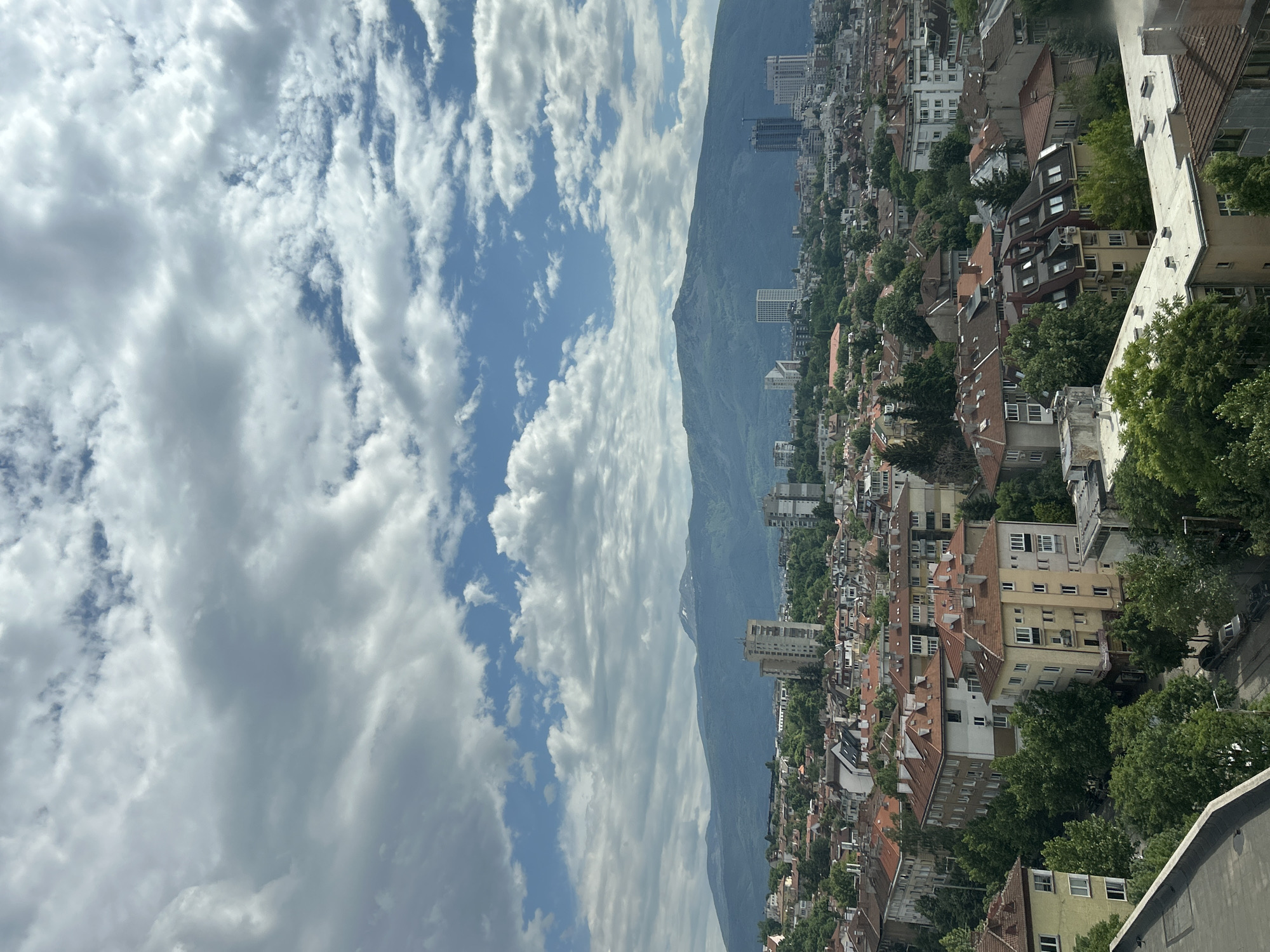 This is a daytime aerial or high-vantage point view of a city, likely Sofia, Bulgaria, set against a prominent mountain range. The image, originally in portrait orientation, captures a vast sky filled with scattered white cumulus and cirrus clouds, with patches of clear blue visible.
In the mid-ground, the imposing Vitosha mountain dominates the horizon, appearing dark green to bluish-grey, indicating distance and lush vegetation. Along the base and slopes of the mountain, the city unfolds, characterized by a dense collection of buildings. Most visible structures are residential, featuring red-tiled roofs, often with multiple stories, and a mix of older and newer architectural styles. Interspersed among the houses are numerous trees, particularly evident in the foreground and lower slopes of the city, suggesting a green and leafy urban environment.
Several taller, more modern multi-story buildings, possibly commercial or apartment blocks, can be seen rising above the general residential sprawl, particularly closer to the mountain base and within the cityscape. No specific activities or people are discernible, and no visible text is present. The overall impression is one of a vibrant city nestled under a grand natural landmark on a bright, partly cloudy day.