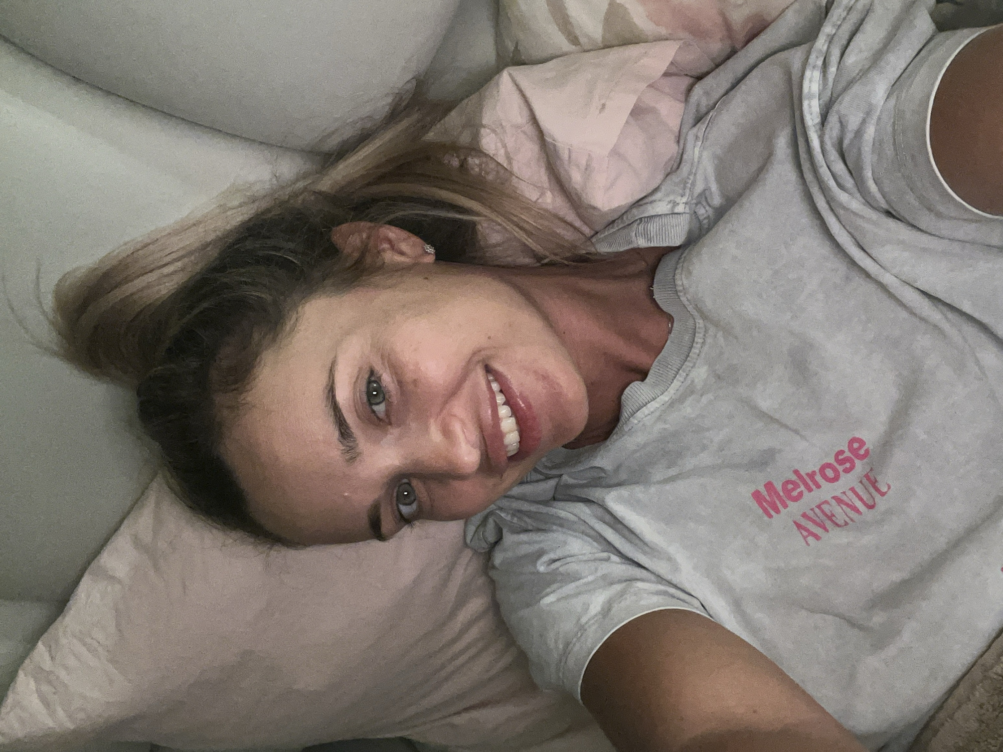 A woman is lying on a bed with pink and white pillows. She is wearing a grey t-shirt with "Melrose Avenue" printed in pink. Her hair is pulled up in a ponytail. She is smiling at the camera. The lighting suggests it is nighttime, and the setting appears to be indoors. No specific location in Sharm El Sheikh, Egypt can be determined from the image.