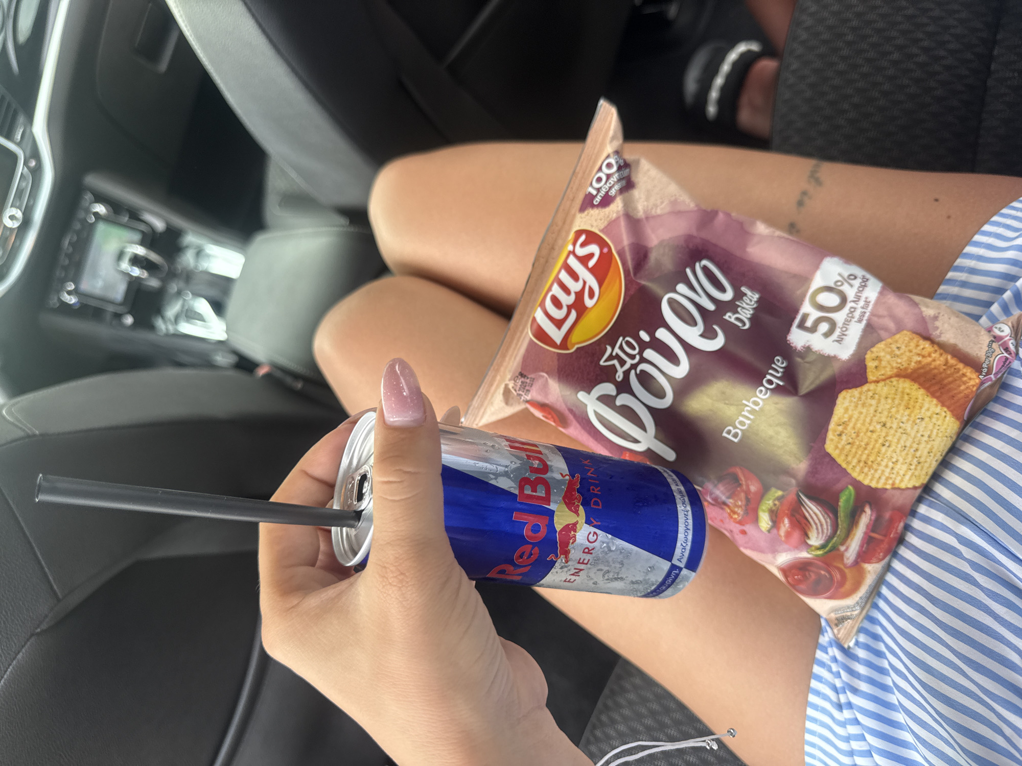 A woman is sitting in a car and holding a can of Red Bull with a straw in it in her left hand. She is also holding a bag of Lay's potato chips (Barbeque flavor) in her right hand. The woman is wearing a blue and white striped shirt. The interior of the car is visible in the background. The location is undetermined.