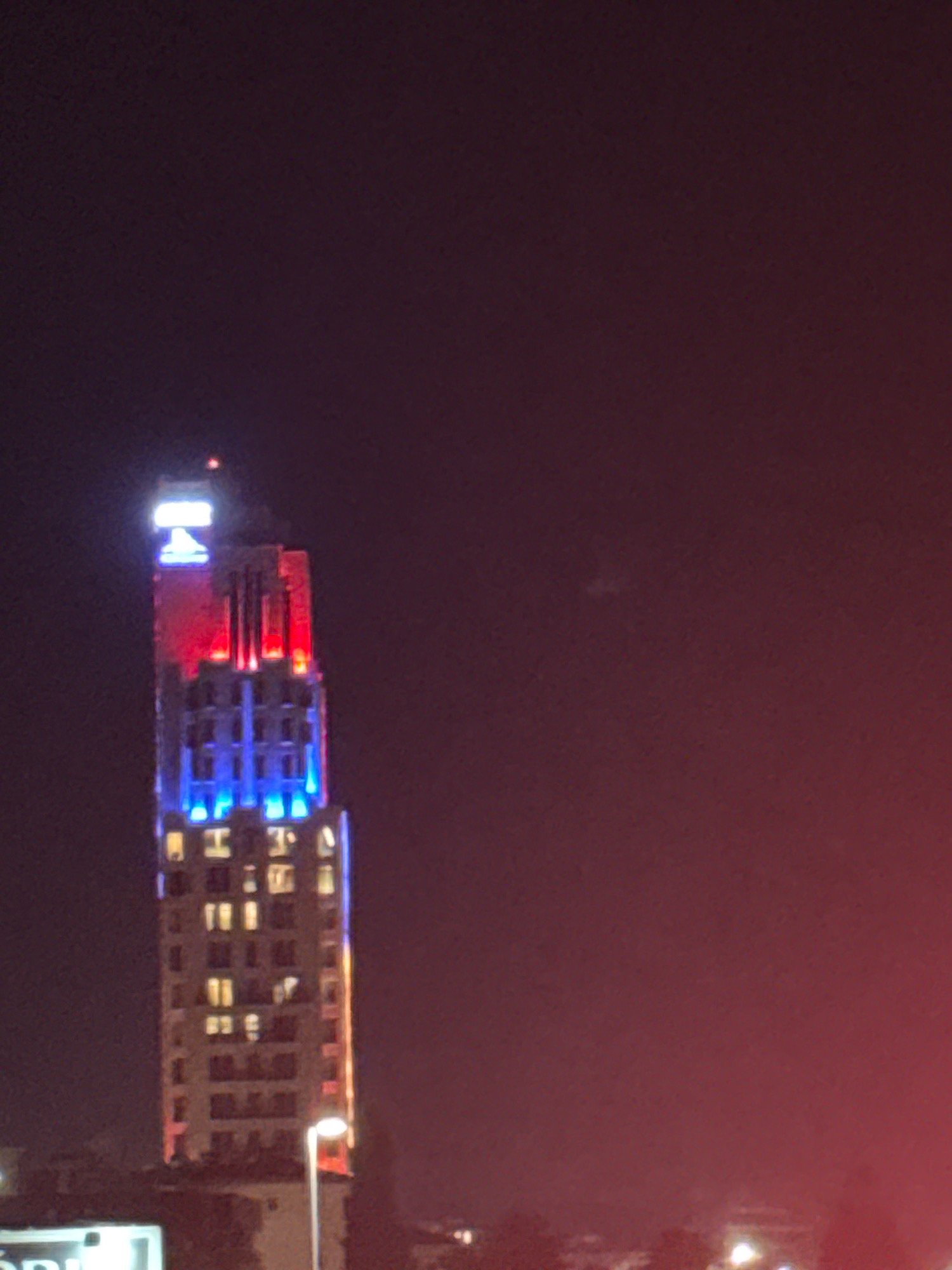 A night time, medium shot captures a tall, slender building illuminated by red, white, and blue lights. The building's upper section is highlighted with bright white and blue lighting, topped with a neon "H" sign. The middle section is bathed in red, and the lower section in blue. Numerous windows on the building's facade are lit with a warm yellow glow, indicating occupancy. A streetlamp with a bright orange light stands in the foreground to the right of the building. The overall scene is dark, with a reddish hue in the background, suggesting ambient light or a hazy atmosphere. The location is identified as Agios Tychona Community, Cyprus.