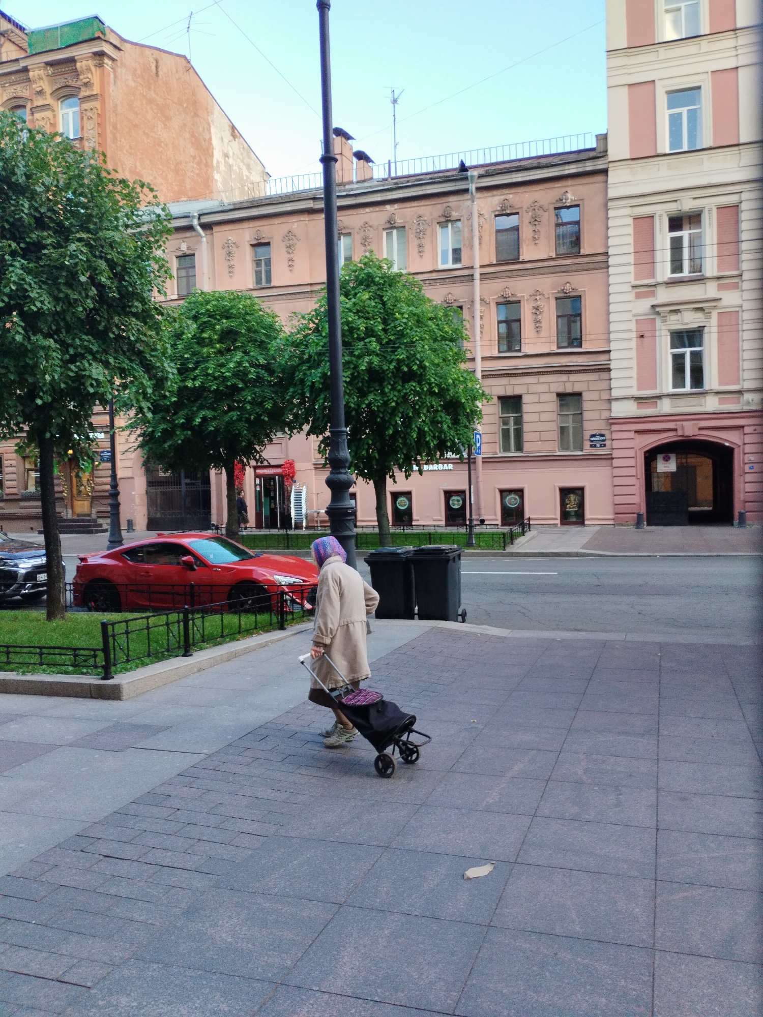 The image depicts an exterior scene in Saint Petersburg, Russia, with a focus on a street and the facades of buildings. A woman, wearing a headscarf and a beige coat, is seen from the back, pulling a wheeled shopping bag. She is walking on a sidewalk paved with square tiles. Adjacent to the sidewalk is a patch of grass enclosed by a black metal fence, beyond which sits a red sports car. Behind the car are trees, and further back are buildings with pink and orange facades, featuring multiple windows and decorative architectural elements. A tall, dark lamppost stands on the sidewalk. The sky is overcast.