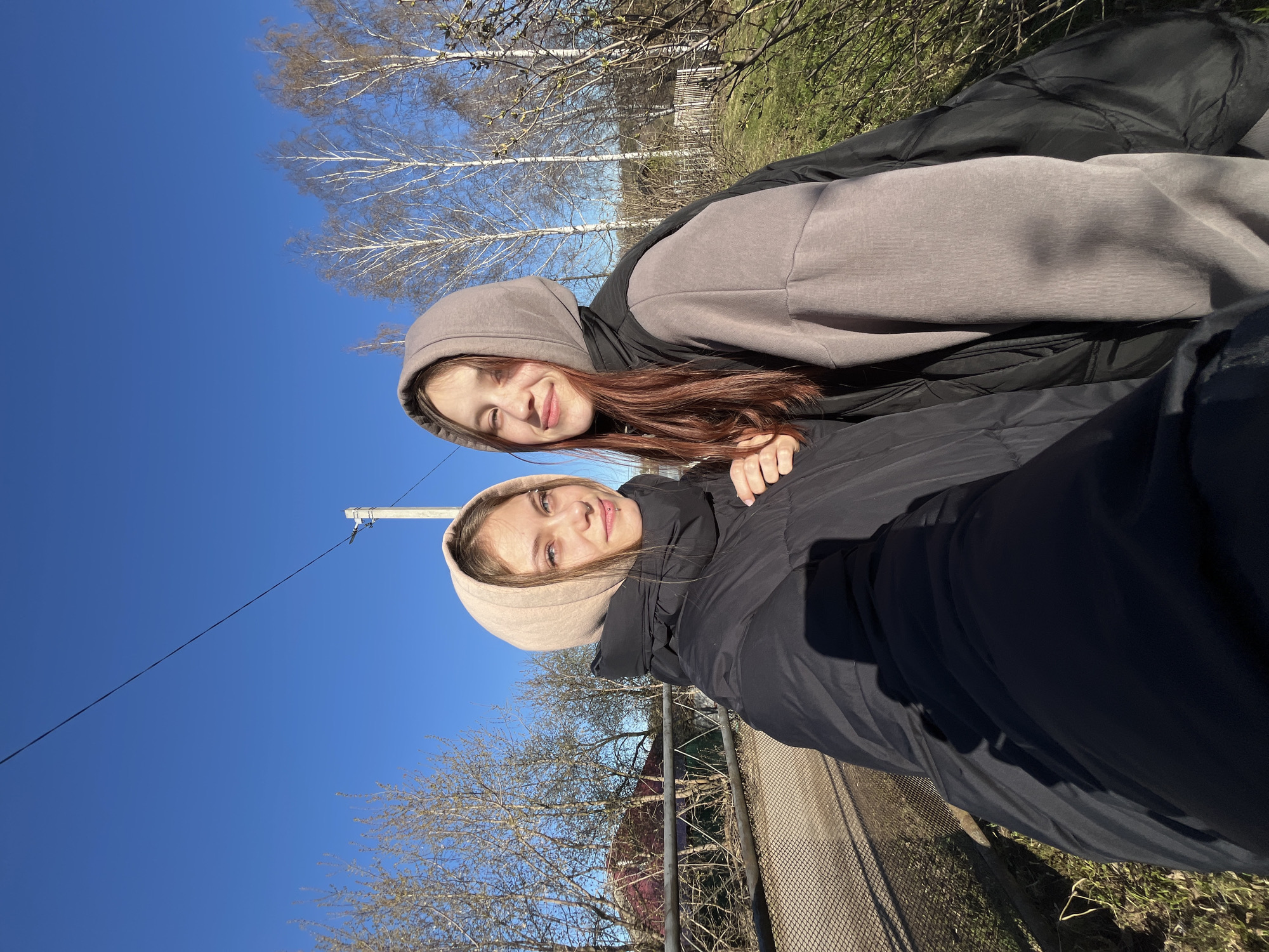 This image captures two young women posing for a selfie outdoors on a bright, sunny day in Фоки, Russia. The photograph is originally rotated 90 degrees counter-clockwise, with the sky appearing on the left and the ground on the right.
If viewed in its natural orientation, the clear blue sky would occupy the upper portion of the frame, and the ground the lower. The woman positioned closer to the camera, on the left, has light brown hair and is wearing a light beige hoodie beneath a black puffer jacket. She has noticeable lip and ear piercings and offers a subtle smile directly at the camera. Her left hand rests gently on the arm of the other individual. The second woman, standing slightly behind and to the right, has reddish-brown hair and is dressed in a dark grey hoodie under a black puffer jacket. She displays a broad smile, also looking at the camera.
The background showcases a vast, unblemished blue sky. Several deciduous trees with bare, pale branches, resembling birches, are prominent against the sky, suggesting either late winter or early spring. The ground beneath them is covered with dry, light-colored grass mixed with nascent green patches. A chain-link fence runs horizontally across the middle ground. A utility pole with a single wire is visible towards the upper left section of the frame. The overall atmosphere conveyed is cheerful and relaxed, with both subjects appearing happy under the clear, bright daylight. No visible text is present in the image.