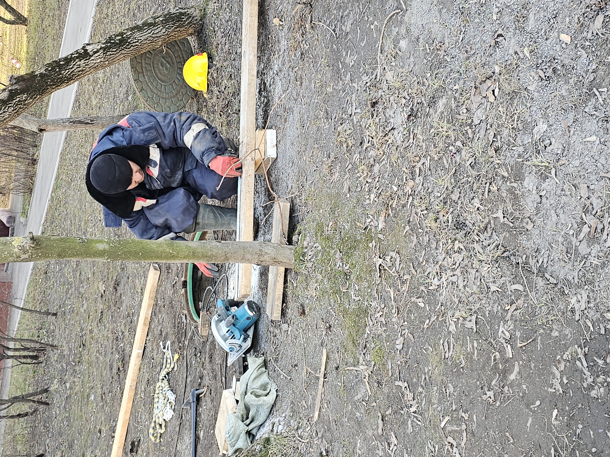 A man in a blue and gray work suit, black hat, red gloves, and green boots is working outdoors. He is crouched over a wooden structure he appears to be assembling. He is connecting wood pieces with string or twine. A yellow hard hat is on the ground nearby. A circular saw, pieces of wood, a chain with hooks, a clamp, and a green cloth are also on the ground. The work area appears to be a yard or unpaved area with trees. It may be in Moscow, Russia.
