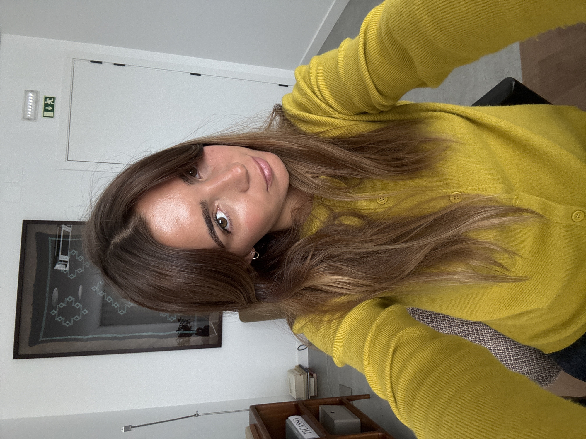 A woman with brown hair and a yellow cardigan is in an indoor setting. A framed picture hangs on the white wall. A wooden table is located below the picture with some books and objects placed on it. An exit sign is located at the top left on the wall above the picture. The scene is potentially located in a residential or office space, though there is insufficient information to verify its location in Torres Vedras, Portugal.