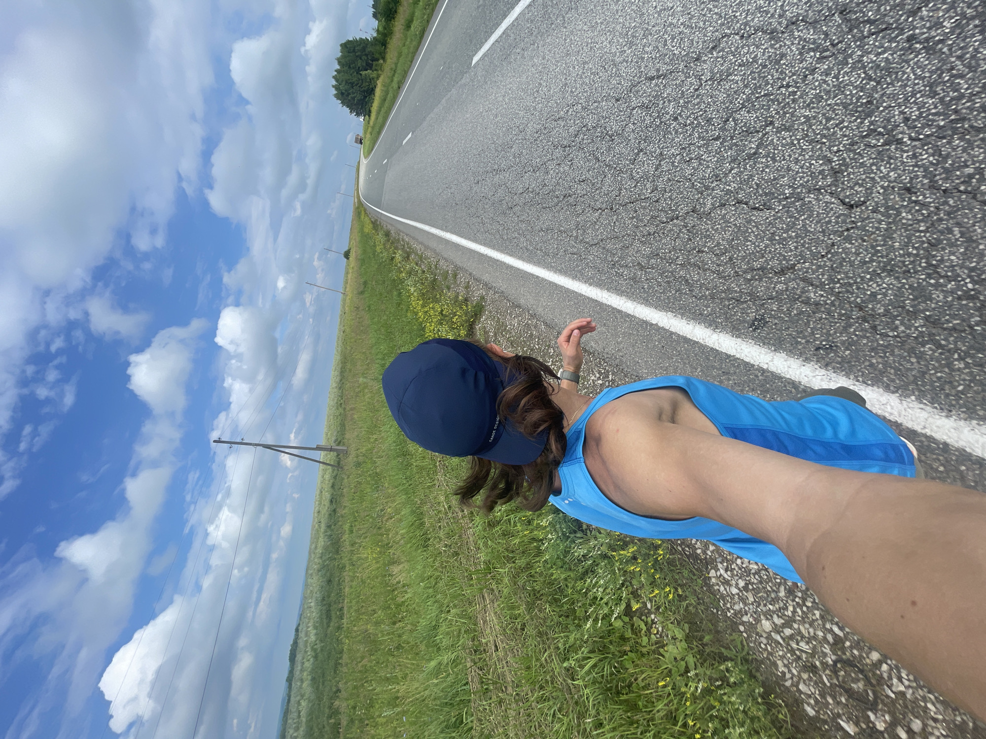 An individual wearing a blue tank top and a dark blue baseball cap is running on the shoulder of a paved road. The road is grey and appears to have some cracks. The individual's right arm is extended forward, holding a camera, creating a selfie perspective. The individual's hair is brown and appears windblown. The background consists of a lush green field with some yellow wildflowers on the left and the open road stretching into the distance on the right. There are utility poles with power lines running along the side of the road. In the distance, trees and a truck can be seen. The sky is blue with scattered white clouds. The scene appears to be in Perevoznoe, Russia.