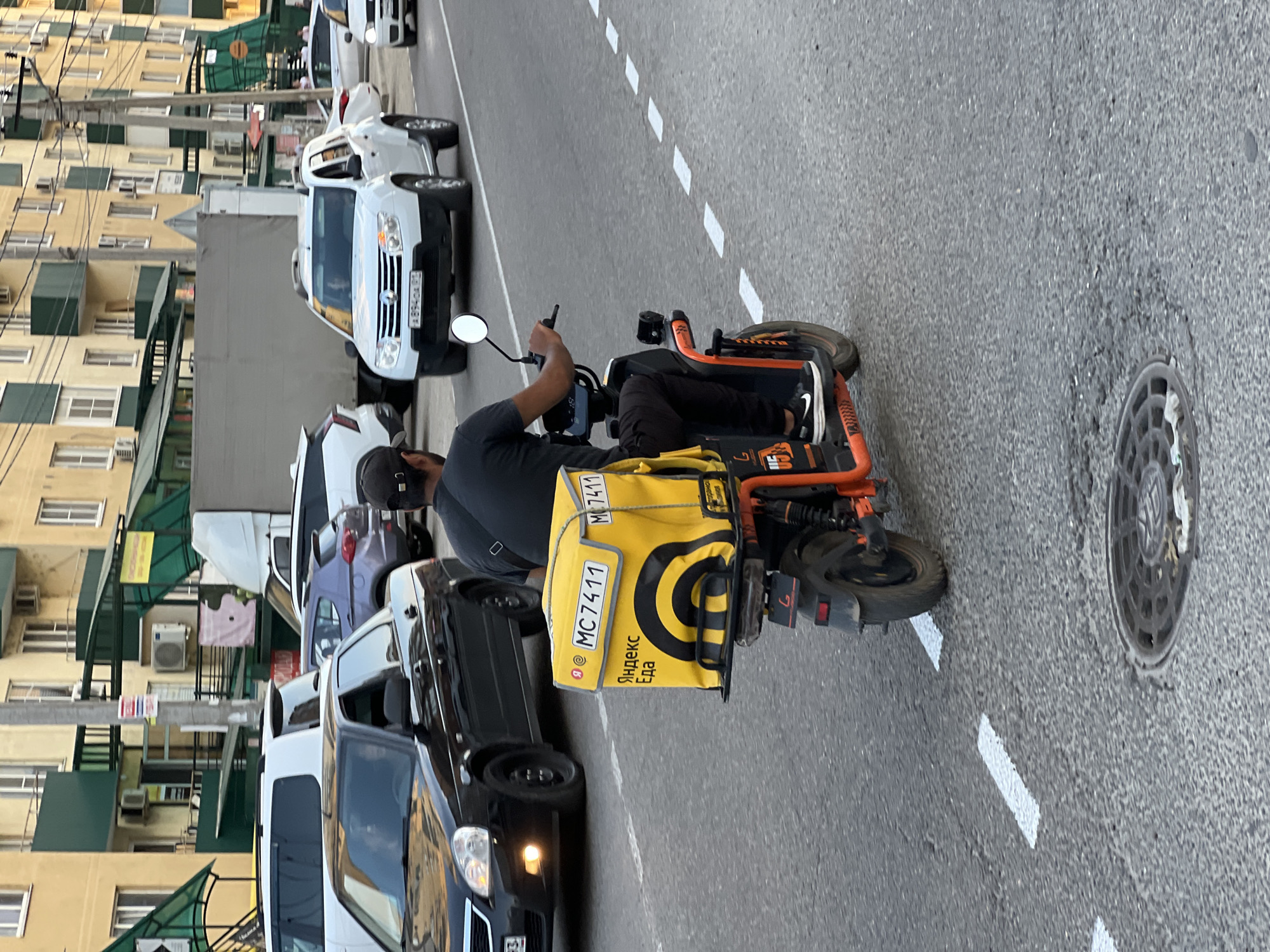 An adult male wearing a black t-shirt, black pants, and black Nike sneakers is riding an orange electric scooter down a paved road in Yablonovskiy, Russia. The scooter has a large, yellow insulated delivery bag attached to the rear, which displays the Yandex.Eda logo and the text "MC 7411" in black lettering. The man is looking forward, holding onto the handlebars. A black sedan is in the lane to the left of the scooter. Further ahead, a white SUV is in the right lane, and other vehicles are visible in the background. Buildings line the street on the left, with shops and signs visible. The road has white dashed lane markings and a manhole cover is present in the foreground. The lighting suggests it is daytime, possibly late afternoon or early evening.