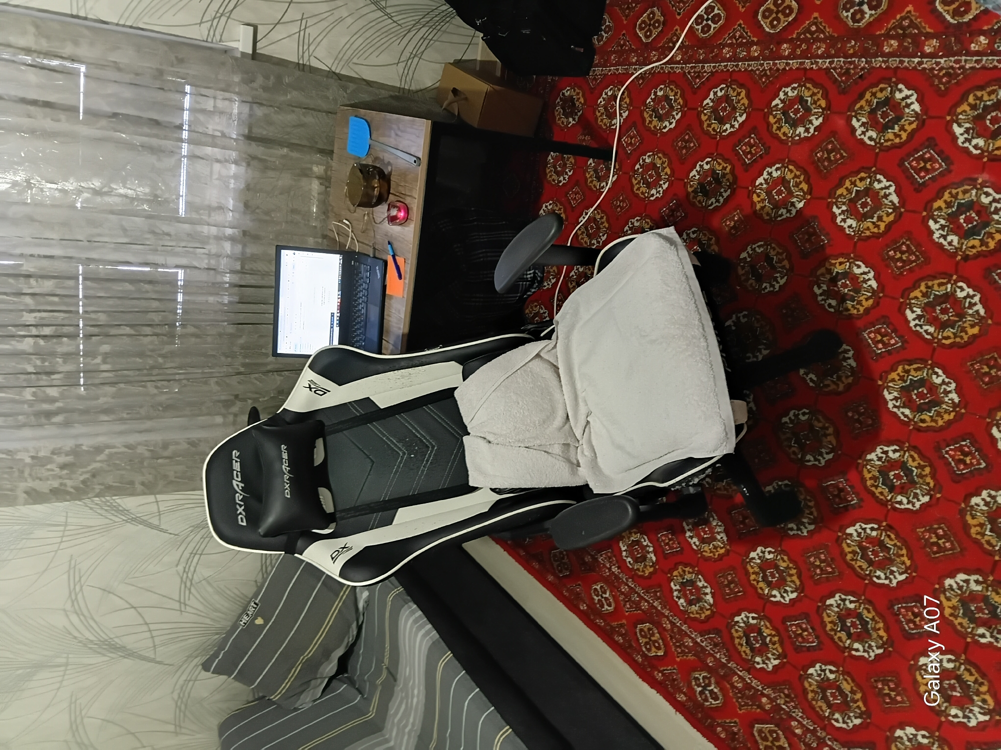 The image shows a gaming chair with a white towel draped over it, positioned on a red carpet with a traditional pattern. A laptop is open on a desk to the left of the chair, with papers and a pen visible on the desk. To the right of the laptop, a blue object, possibly a spatula, and a round metal container are present. The setting appears to be a room with a window covered by sheer blinds to the left, allowing diffuse light to enter. The overall scene suggests a home office or gaming setup. The carpet's intricate design and the presence of traditional patterns hint at a location in Central Asia, aligning with the provided location context of Tashkent, Uzbekistan. No people are visible in the image. The lighting suggests it could be daytime, but no specific time can be determined. The text "DXRACER" is visible on the chair multiple times, and "Galaxy A07" is faintly visible on the carpet.