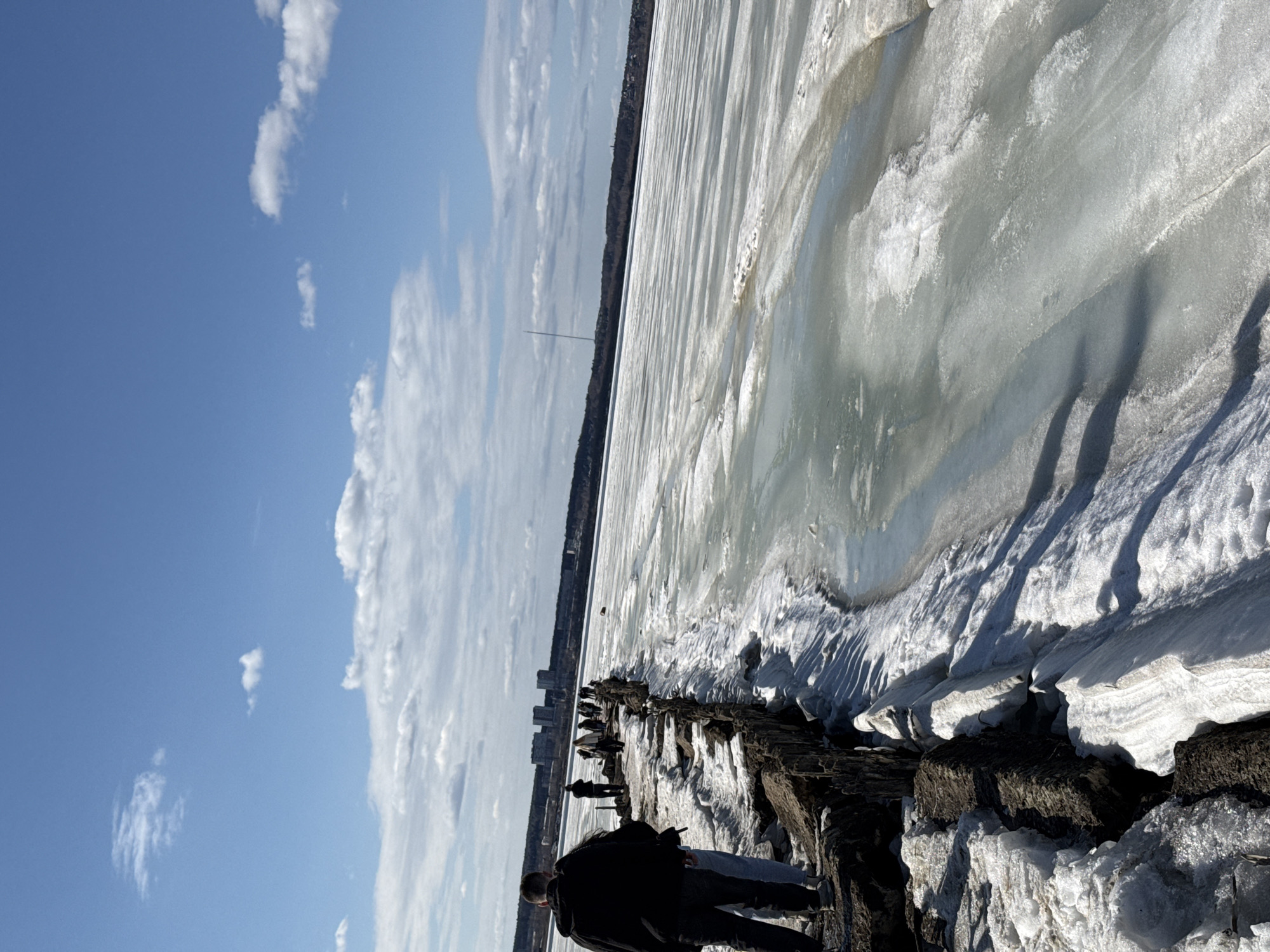 The image depicts a partially frozen body of water, likely a lake or river, on a sunny day with a blue sky and scattered clouds. Along the shoreline, there is a person standing on a rocky or wooden structure. In the distance, a city skyline is visible. The image appears to be taken in a cold climate, as there is ice and snow along the water's edge. Given the location in Izhevsk, Russia, it is probable that the frozen body of water is the Izhevsk Reservoir, also known as Izhevsk Pond, which is a prominent feature in the city.
