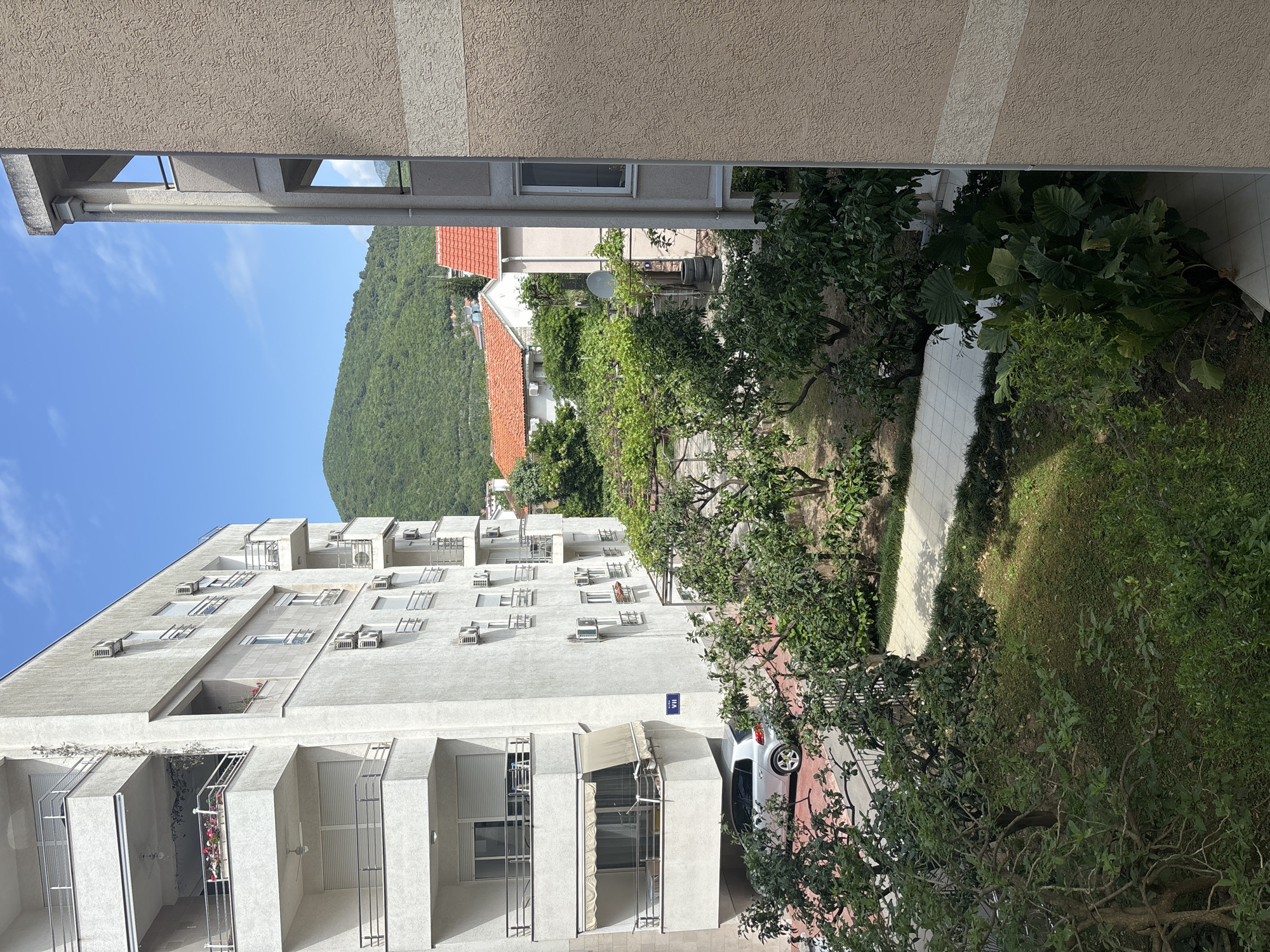 This is an elevated view in Budva, Montenegro, showing buildings, vegetation, and a hillside.
A multi-story white building with numerous balconies and air conditioning units dominates the left of the frame. The building's facade has a modern architectural style.
To the right of this building is a view into a garden with trees, grass, and a white-tiled walkway. A white car is parked on a red surface near the building. Beyond the garden, a hillside covered in green vegetation is visible.
The top of the frame is partially obscured by the top and adjacent building. A clear blue sky is visible between the buildings on the left.
Overall, the image depicts a residential area in Budva, characterized by modern architecture and lush green spaces.
