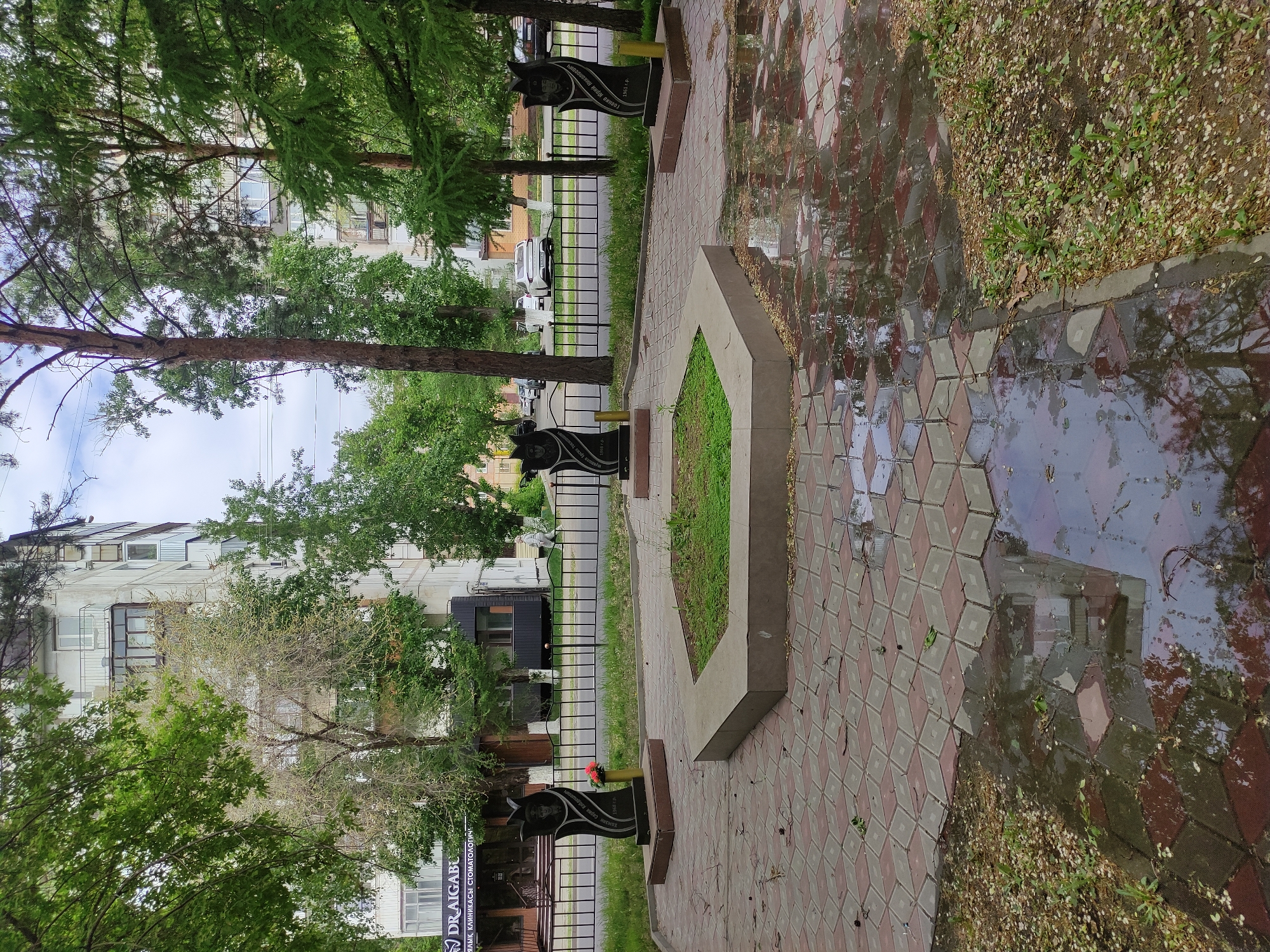 The image depicts a memorial site in Pavlodar, Kazakhstan, likely dedicated to individuals of local significance. The foreground features a brick-paved path, wet from recent rain, reflecting the surroundings. To the left, a small concrete enclosure contains grass. Along the path, several dark stone monuments stand behind a black metal fence. The monuments appear to be engraved with images and text, possibly names and dates, some adorned with flowers. In the background, buildings and trees line the street. A vehicle is visible parked on the side of the road beyond the fence. The overall atmosphere is somber and reflective.