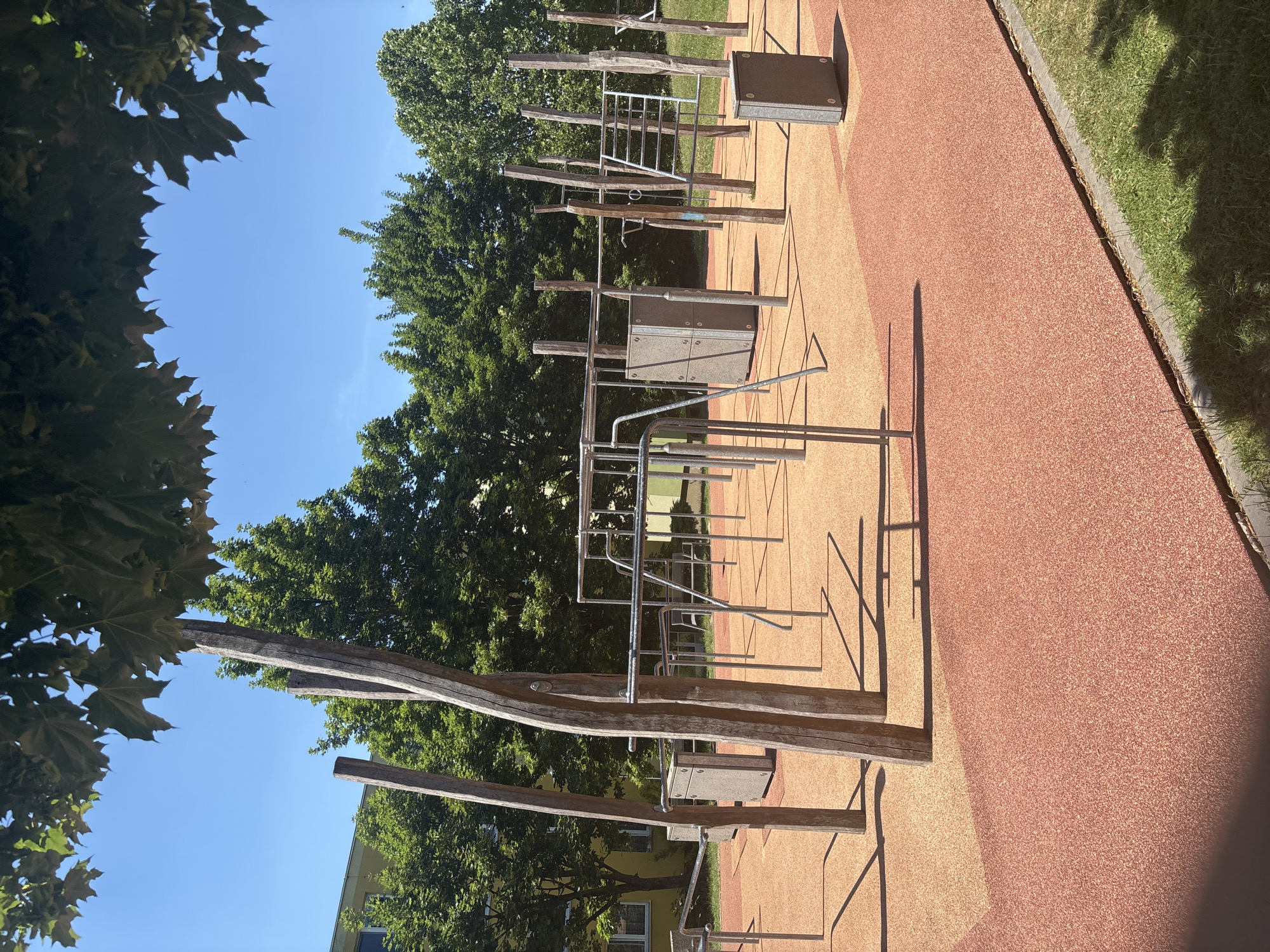 An outdoor fitness park is depicted on a sunny day. The park features an orange rubberized safety surface, with a darker red strip bordering the foreground. Several sets of exercise equipment, made of metal bars and wooden posts, are arranged across the area. These include parallel bars, pull-up bars, and what appear to be agility ladders. Two grey, cube-shaped structures are also visible, one in the mid-ground and another to the right. Behind the fitness equipment, trees form a dense green backdrop, and a light yellow building with windows is partially visible through the foliage. The sky is a clear, bright blue.