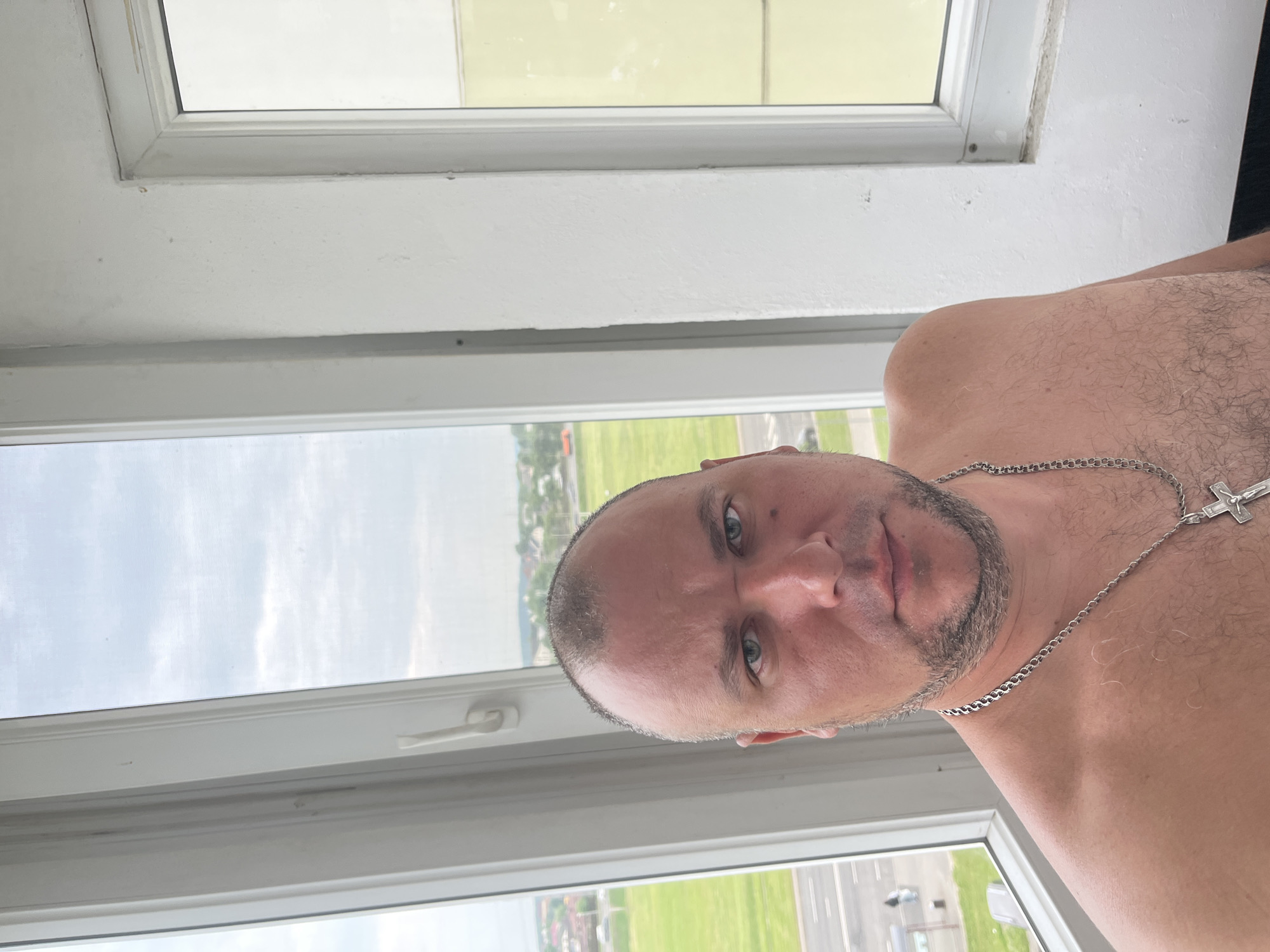 The image captures a close-up of a shirtless man, seemingly a selfie taken indoors next to a window. The man looks directly at the camera with a neutral expression. He has a bald or very closely shaven head and a short, dark beard or stubble. Around his neck, he wears a silver chain with a crucifix pendant.
Behind the man, a large white window opens to an outdoor scene bathed in daylight. Through the window, a wide expanse of green grass is visible, alongside what appears to be paved sections resembling an airfield runway or large industrial area. In the further distance, faint buildings and trees line the horizon under a partly cloudy sky. The white window frame shows some signs of wear and a handle is visible on one of the panes. The time of day is clearly daytime, with bright ambient light. No text is visible in the image. The provided context indicates the location is Minsk, Belarus.