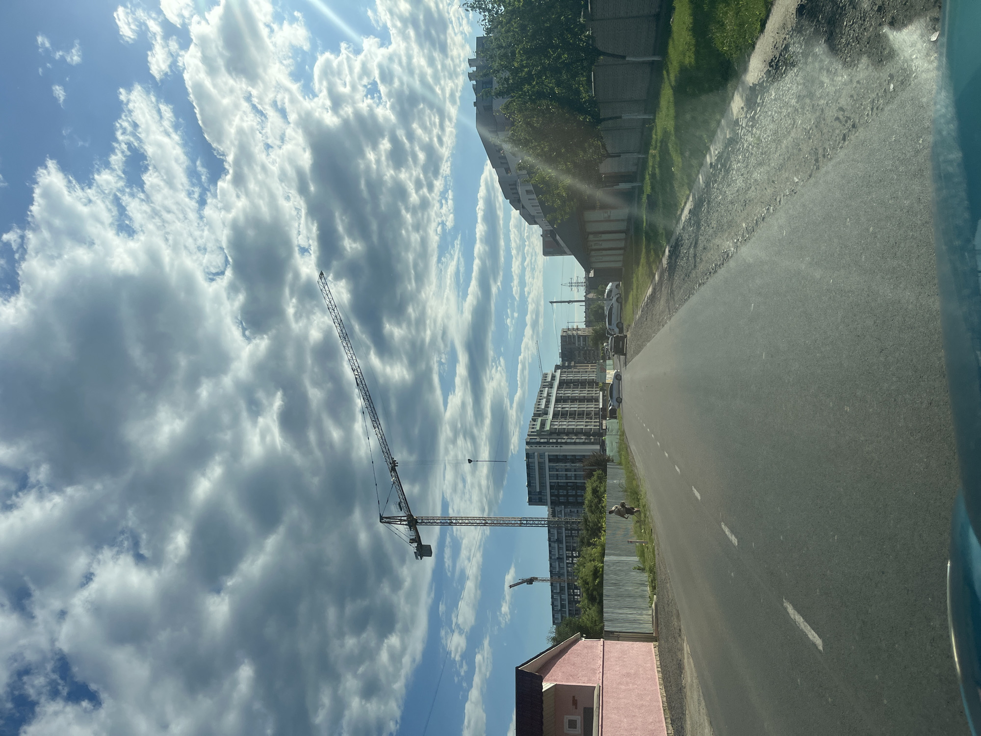 The image depicts a street scene in the city of Storozhnytsia, Ukraine, under a partly cloudy sky. In the foreground, a paved road with white dashed lines runs towards the horizon. On the left side of the road, a pink building with a dark roof is visible behind a corrugated metal fence. Further down the road, a lone individual, appearing to be a male wearing dark shorts and a light-colored shirt, walks along the grassy verge. Several cars are parked or driving on the road. In the background, large multi-story buildings are under construction, with prominent tower cranes dominating the skyline. One large crane is positioned in the center of the frame, its jib extending horizontally across the sky. Another smaller crane is visible to the left. Trees and vegetation line the right side of the road, behind a fence. The overall impression is one of ongoing development and urban expansion.