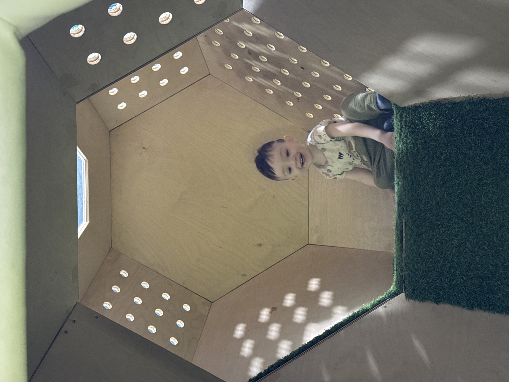 This low-angle photograph captures a young child smiling brightly inside a geometrically shaped play structure in Izhevsk, Russia. The structure is primarily constructed from light-colored wood panels, possibly plywood, forming hexagonal or octagonal sections.
The main subject is a happy toddler or preschooler with short dark hair, wearing a light-colored short-sleeved top with a subtle pattern and olive green pants. The child is positioned towards the right side of the frame, looking up and directly at the camera with a wide, joyful smile. They appear to be sitting or kneeling on a surface that transitions from the wooden paneling to a patch of dark green artificial grass.
The interior walls and ceiling of the structure feature numerous small, circular cutouts. Bright sunlight streams through these holes, creating distinctive circular patterns of light and shadow across the wooden surfaces and the artificial grass. Some of the holes reveal glimpses of a bright blue sky or light source outside. A larger, rectangular opening, resembling a small window, is visible on the left side of the structure, also admitting ambient light.
The scene conveys a playful and bright atmosphere, indicative of an indoor play area or interactive exhibit. The time of day appears to be sunny, daytime, given the strong natural light filtering into the structure. There is no visible text in the image.