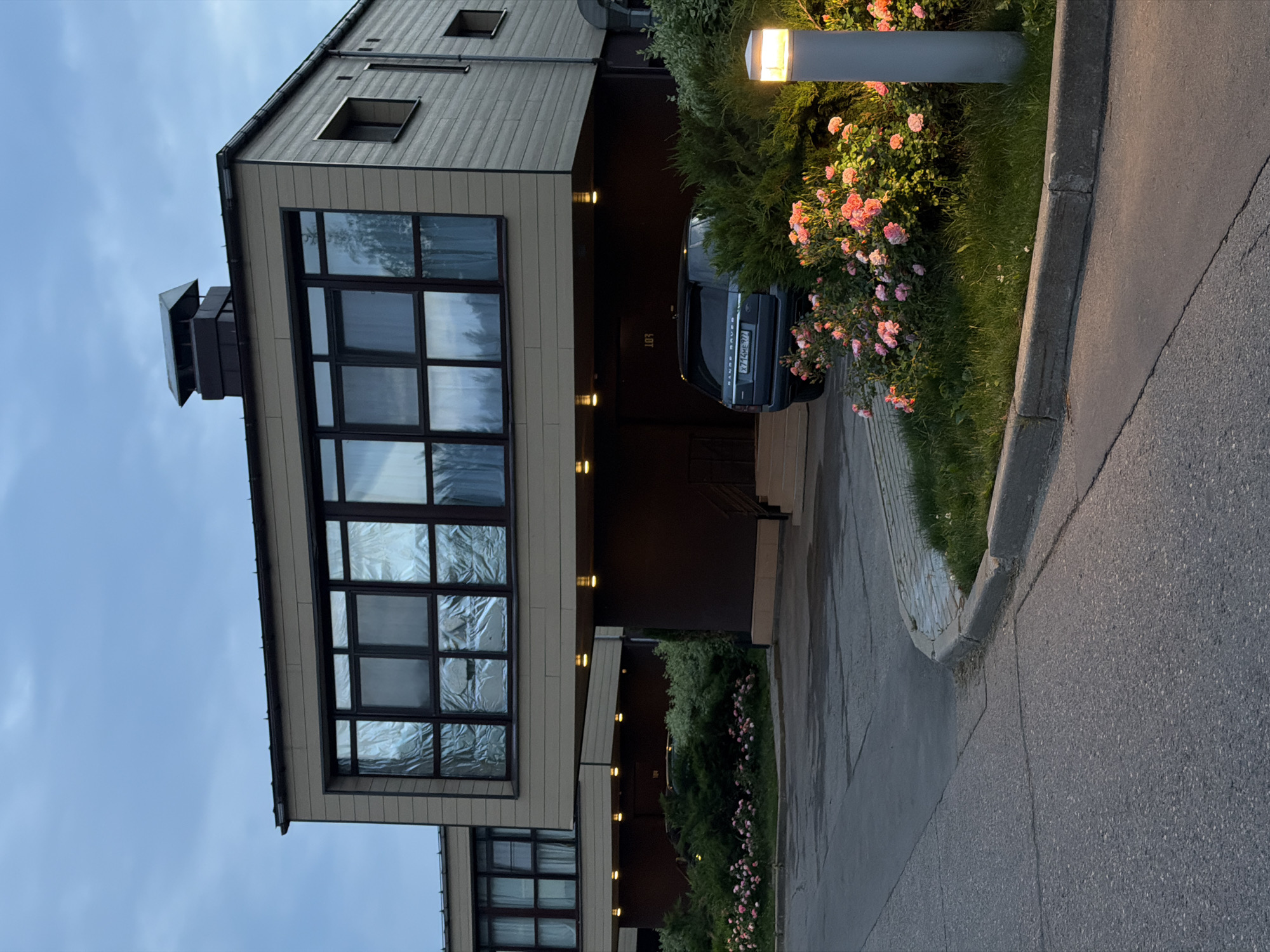 The image shows a modern, multi-story residential building in Skolkovo, Russia, with a contemporary architectural style featuring clean lines and geometric shapes. The building has light-colored siding and dark-trimmed windows. In the foreground, a grey Range Rover is parked in front of the building, partially obscured by a flowering bush with pink roses. A paved driveway leads to the building, and a contemporary lamp post illuminates the area. The sky is overcast and the overall lighting suggests it is dusk or early morning. The environment appears to be a well-maintained residential complex with landscaping.