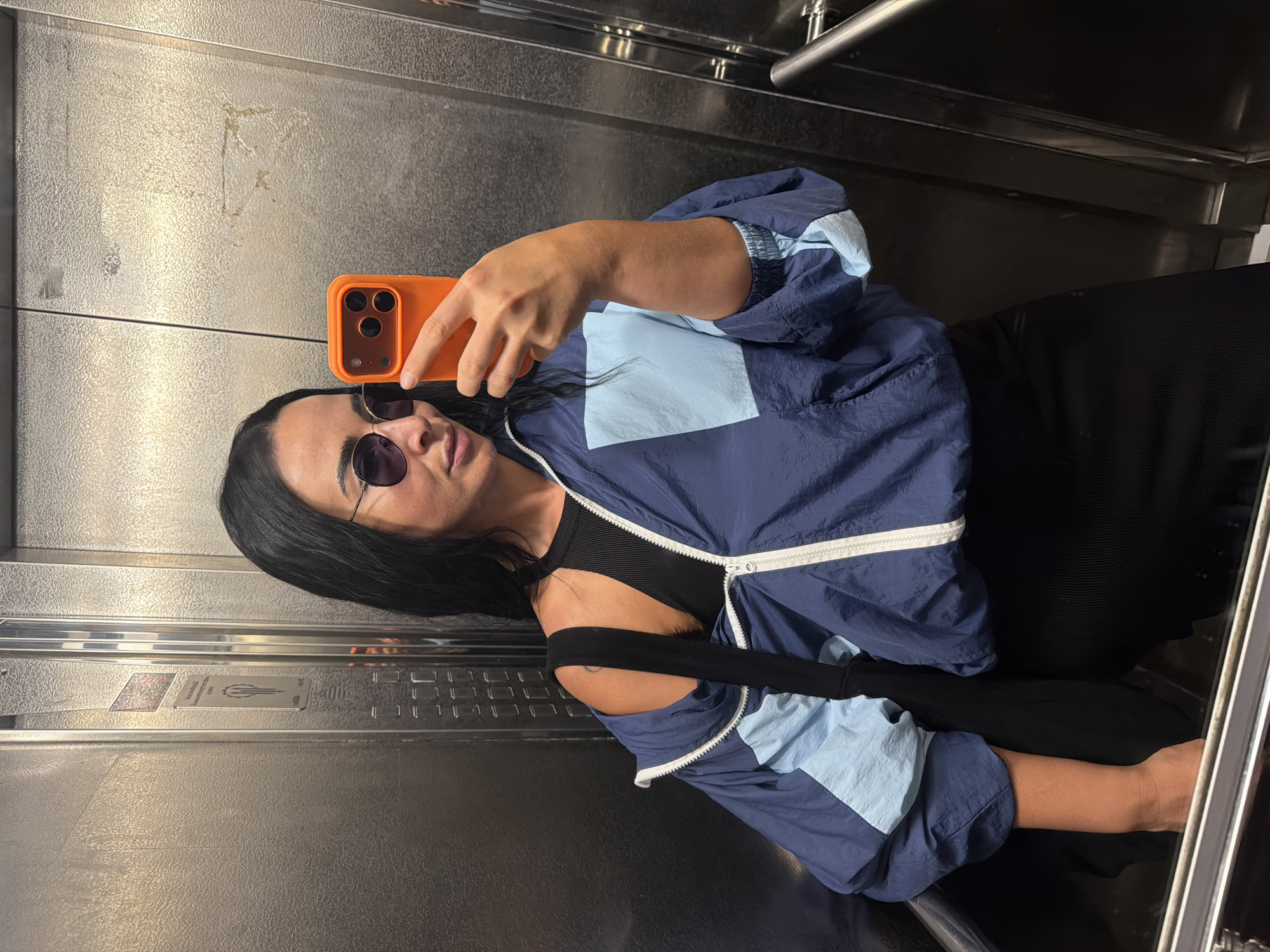 A person is taking a mirror selfie in an elevator. The person has dark hair and is wearing sunglasses, a black top, and a blue and light blue color-blocked windbreaker jacket. They are holding an orange smartphone up to take the picture. The elevator walls appear to be made of brushed metal, reflecting the scene. The lighting is consistent with indoor lighting. The overall mood appears casual. There is visible text on a panel inside the elevator, but it is too blurry to read clearly.