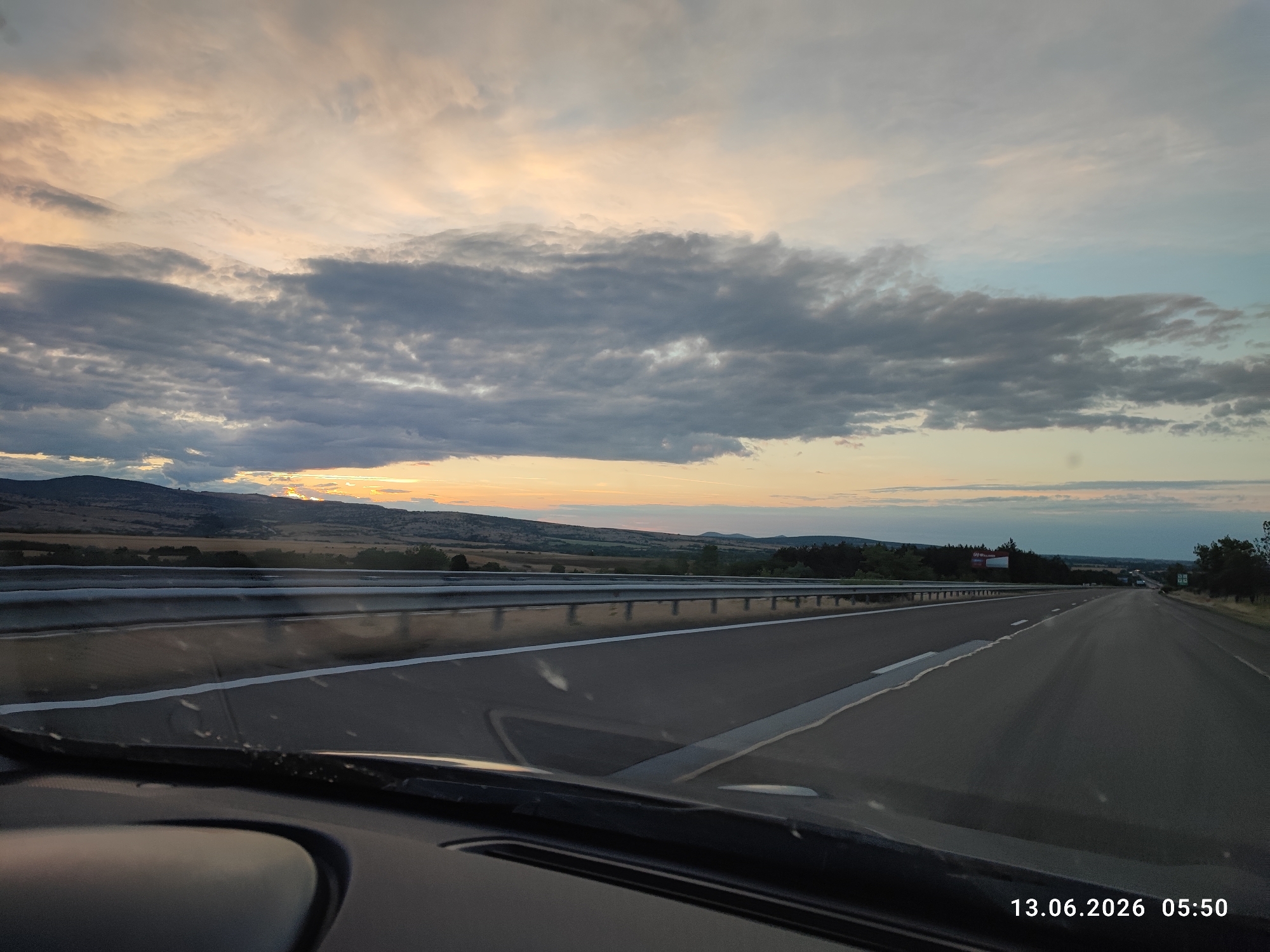 The image shows a view from inside a car, looking out at a highway at dawn. The sky is filled with clouds in shades of gray, white, and pale orange, suggesting the sun is low on the horizon. The highway stretches ahead, with clear lane markings. On either side of the highway are guardrails and a landscape of rolling hills and scattered trees. In the distance, the hills are dark against the brighter sky. The foreground shows the car's dashboard and windshield, with some water droplets on the glass. The date and time stamp in the lower right corner indicates "13.06.2026 05:50". The location of Kalugerovo, Bulgaria, is not explicitly visible or discernible from the image itself.