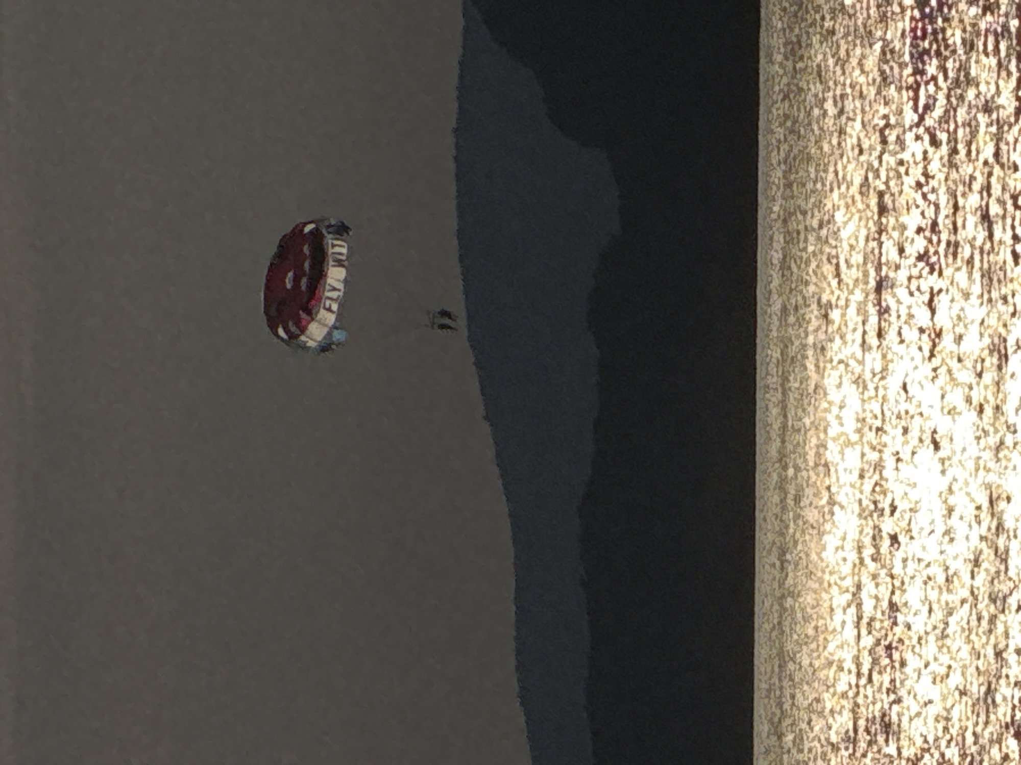 This is a photograph of a parasailer in the air over the water. The parasail is red and white with the words "FLY WITH" visible on it. A person can be seen attached to the parasail. The scene is set near the coast, with dark water and a textured, bright surface on the right side of the frame, suggesting a sandy beach or shoreline. The lighting implies it is either daytime or a bright evening.