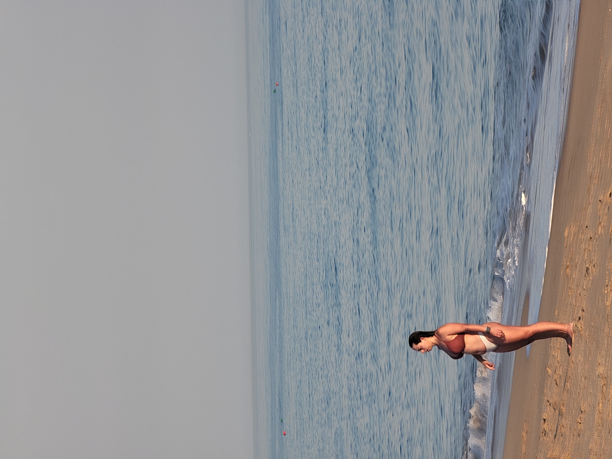 A woman stands on a sandy beach in Quarteira, Portugal, facing left in profile. She is wearing a brown bikini top and white bikini bottoms. Her dark hair is wet and pulled back. The sea is calm with gentle waves lapping the shore. The sky is overcast and a light gray. In the distance, there are a few buoys in the water. The sand is a light brown color and has some footprints.