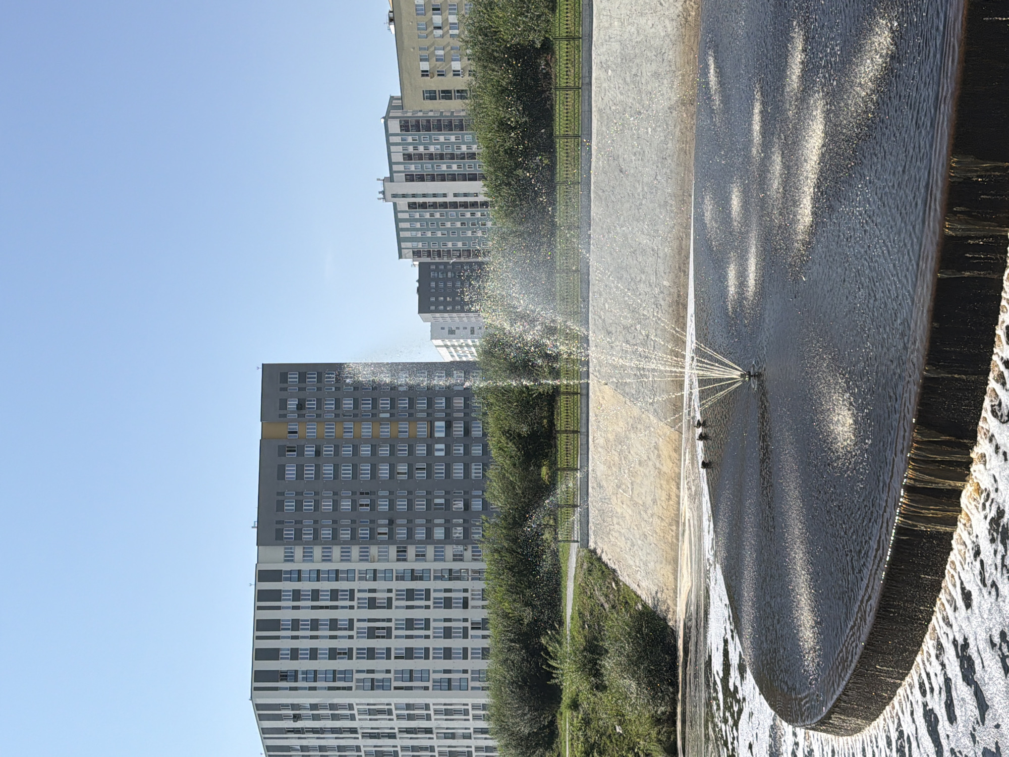 The image shows a modern urban waterfront scene in Yekaterinburg, Russia. In the foreground, a circular fountain with multiple jets of water creates a spray that catches the sunlight. The fountain is situated on a dark, flat surface that appears to be overflowing with water, creating a cascading effect down its edge. Beyond the fountain, a concrete embankment slopes upwards, covered in stone. Behind the embankment is a line of lush green trees and bushes. In the mid-ground, a black metal fence with an ornate pattern runs along the top of the embankment, separating the waterfront from a paved walkway or path. Dominating the background are several multi-story apartment buildings of varying designs and colors, showcasing modern architecture. The sky is a clear, bright blue, indicating a sunny day. The scene appears to be a public park or recreational area within the city.
