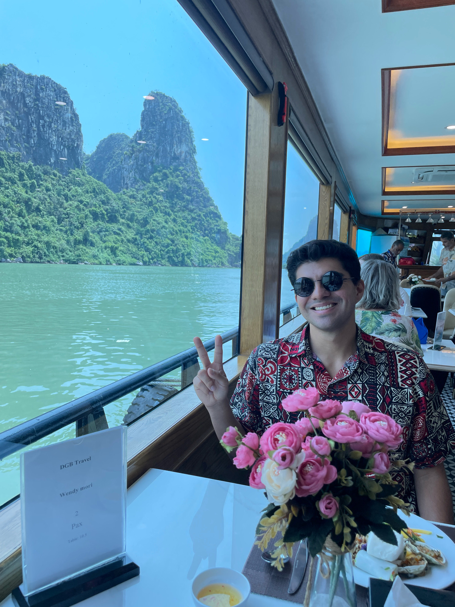 A man is seated at a table on a boat, smiling and making a peace sign with his right hand. He is wearing a patterned short-sleeved shirt and sunglasses. In front of him, a vase holds a bouquet of pink and white flowers. A menu on a stand on the table reads "DGB Travel", "Wendy mori", "2 Pax", and "Table 10.5". The boat is passing by limestone karst formations and green vegetation typical of Ha Long Bay, Quang Ninh City, Vietnam. The water is a greenish-blue. Other diners are visible in the background.