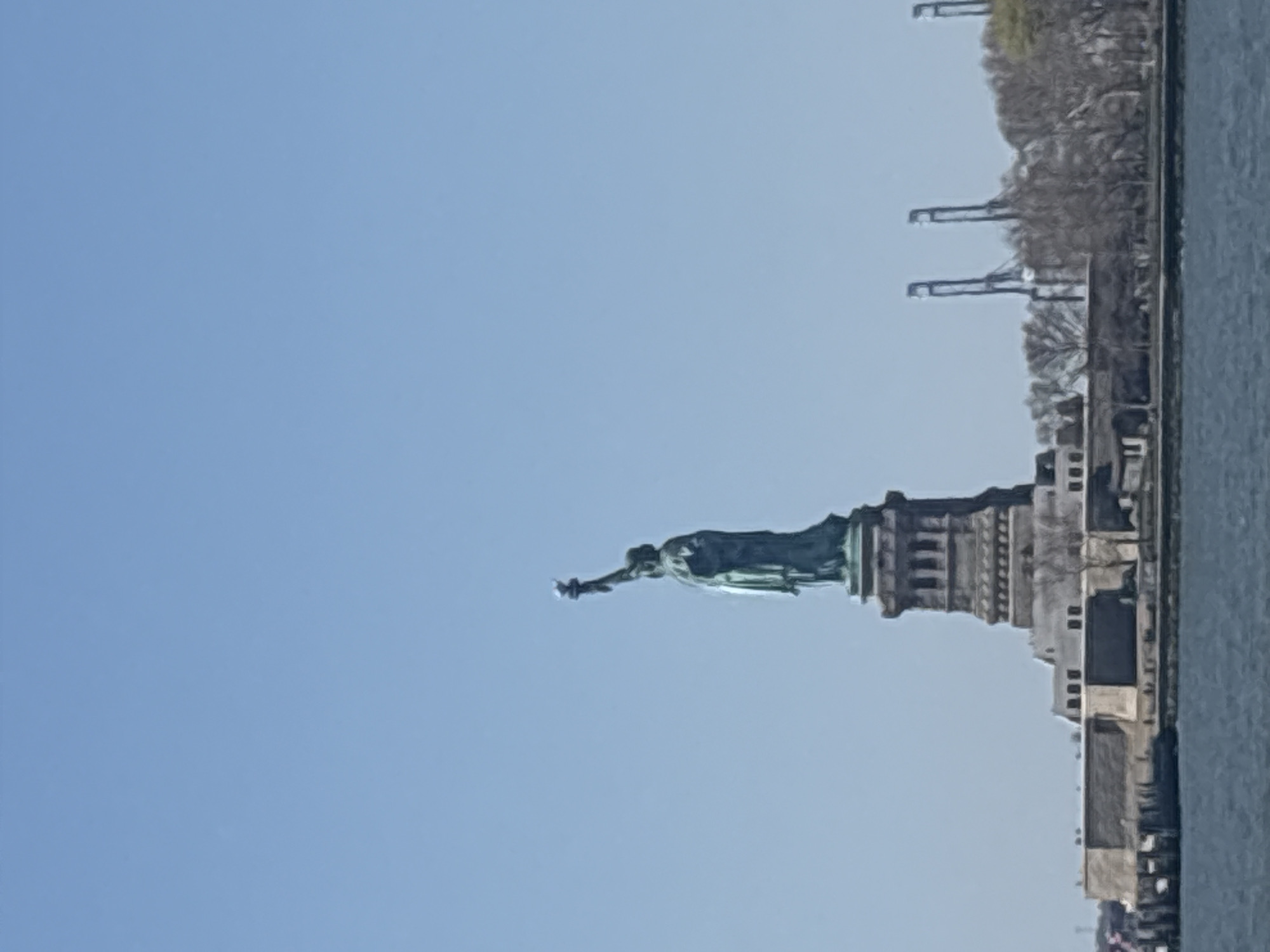 The image shows the Statue of Liberty viewed from the water, likely from a boat. The statue is green and its base is gray. Behind the statue, on the right, are trees, a building, and industrial cranes. The sky is clear and blue. The water is dark gray.