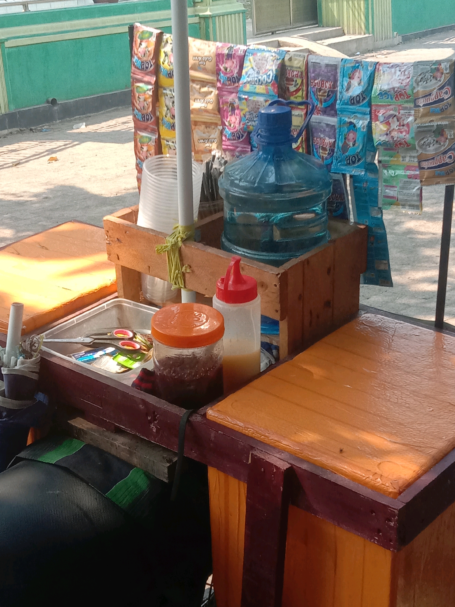 The image depicts an outdoor scene in Ngempit, Indonesia, likely a street or market stall. In the foreground, a wooden cart is visible, laden with various items. A large blue water cooler bottle sits prominently on the cart, its contents a clear blue liquid. Next to it, a wooden box holds a stack of white disposable cups. A glass jar with an orange lid contains a dark, granular substance, possibly spices or sugar. A clear plastic squeeze bottle with a red lid contains a yellowish liquid, likely a sauce or condiment. A metal tray holds a pair of scissors and several small packets, some appearing to be food items. A dark seat, possibly for a motorcycle or scooter, is partially visible beneath the cart. In the background, a green wall provides a backdrop, with several rows of colorful packaged snacks or candies hanging in front of it. The ground appears to be a dirt or gravel surface, with sunlight casting shadows, suggesting it is daytime.