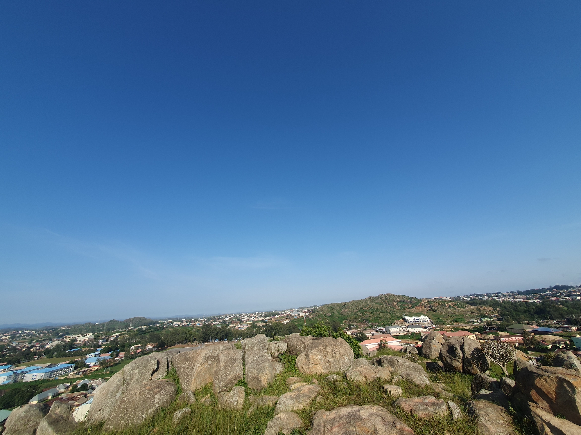 A high-angle panoramic view captures a serene urban landscape in Jos, Nigeria, under a clear, vast blue sky. The foreground is dominated by a cluster of large, weathered rocks covered in sparse green grass, hinting at a natural elevation. Beyond these rocks, the city of Jos unfolds in a tapestry of residential and commercial buildings, interspersed with lush green trees. The architecture is diverse, featuring a mix of modern structures with light-colored roofs and traditional-style houses. A prominent, multi-story building with a white facade and blue accents stands out amongst the other structures. The terrain gently slopes, with hills visible in the distance, adding depth to the scene. The image conveys a sense of tranquility and the harmonious coexistence of nature and urban development.