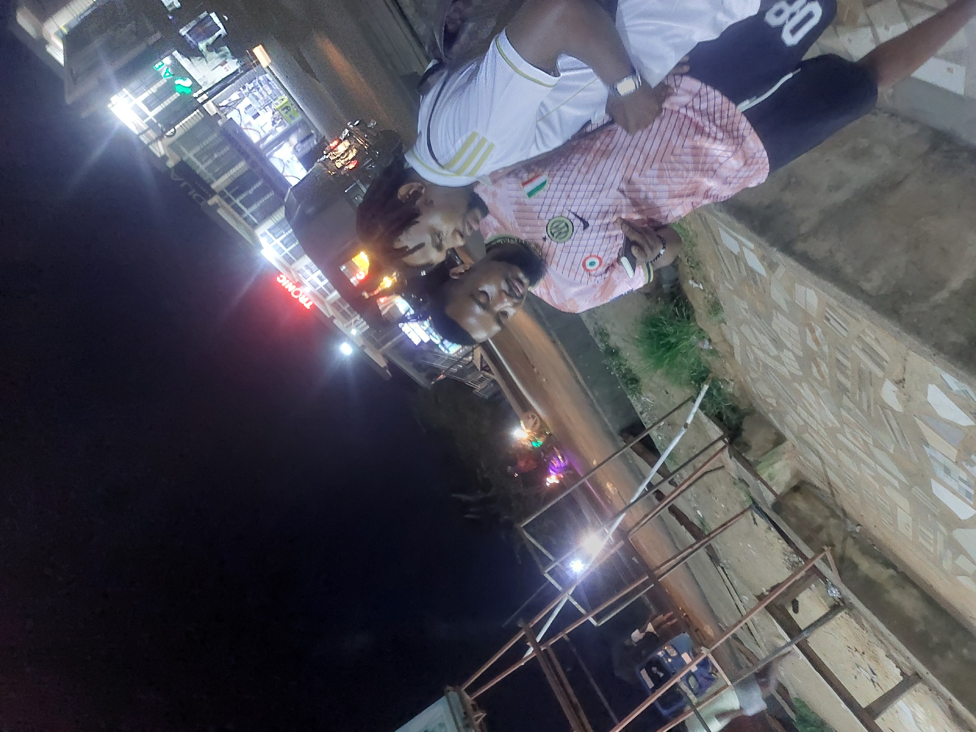 Two men are visible in the image. One wears a white shirt with gold stripes on the shoulder and dark shorts with a white "80" printed on the leg. The other man wears a pink striped shirt and is seen with a necklace.
The background shows a street scene at night. Buildings with lit signs are visible in the distance, including one that reads "TRONIC" in red letters.
The location is likely Dar es-Salaam, Tanzania.