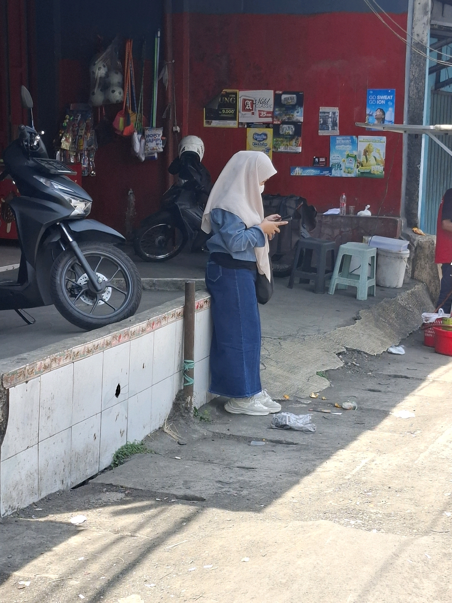 The image depicts an outdoor scene in Cikahuripan, Indonesia, likely on a street or sidewalk in front of a building with a red facade. A young woman wearing a light beige hijab, a blue denim jacket, and a long blue denim skirt stands facing away from the camera, holding a mobile phone in her right hand. She is wearing white sneakers. To her left, a dark-colored scooter is parked. Behind the woman, two stools, one dark green and one light blue, are placed on the ground. Various items are displayed on the red wall of the building, including posters and what appear to be cleaning supplies. In the foreground, the ground is paved with concrete and shows signs of litter, with discarded wrappers and debris scattered around. A drainage channel is visible along the edge of the sidewalk. The lighting suggests it is daytime, with shadows cast on the ground.