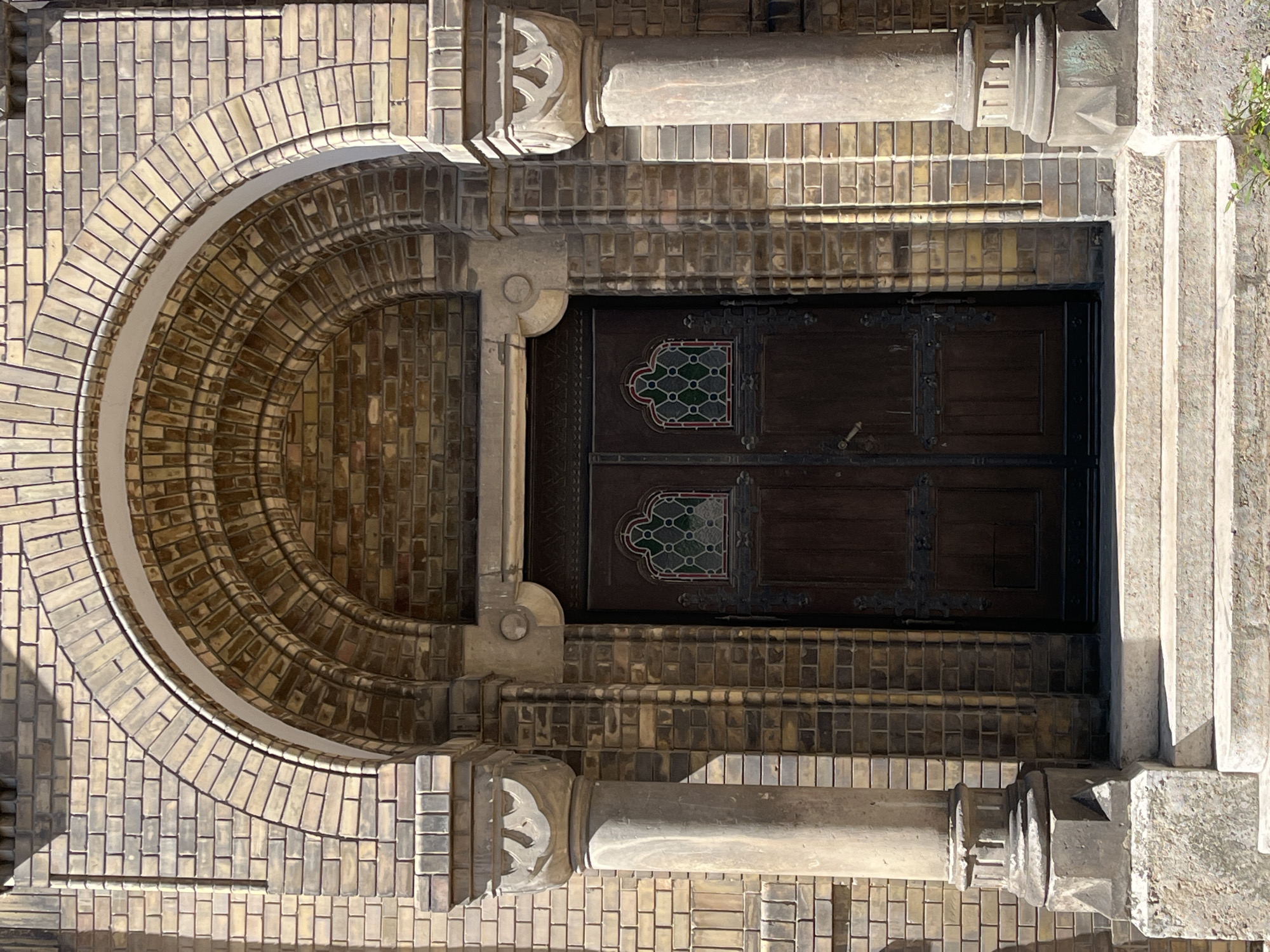 The image captures an ornate architectural entrance, presented from an unusual, rotated overhead perspective. Dominating the right side of the frame is a large, dark brown wooden double door, featuring two distinct rectangular stained-glass panels. These panels are rounded at the top and display intricate geometric patterns in shades of green, white/clear, and red outlines. The door is adorned with heavy, dark metal hardware, including hinges.
The door is set within a rich frame of varied yellowish-beige brickwork, some sections of which exhibit a glazed or iridescent sheen. To the left of the door, a deep, semi-circular archway curves gracefully, constructed from meticulously arranged bricks that create a textured, ribbed effect as it recedes. The archway's inner surface appears darker, suggesting depth and shadow.
Flanking the entrance are two decorative stone elements, resembling truncated columns or pilasters, which appear at the top and bottom of the frame in the current orientation. Each features an intricately carved capital with stylized, interconnected patterns. The surrounding brickwork displays diverse laying patterns, enhancing the visual complexity.
A section of lighter-colored stone steps or a plinth is visible on the far right edge of the image. Strong, crisp shadows are cast by the architectural features, indicating bright, direct sunlight, suggesting a clear day, likely midday. No people or activities are visible. There is no discernible text. The scene conveys a sense of historical grandeur and intricate craftsmanship in Novi Sad, Serbia.