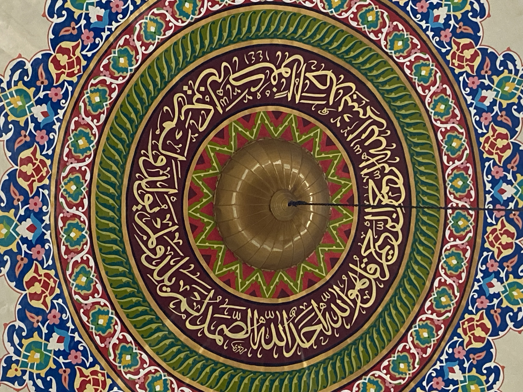 The image shows a close-up, eye-level view of the ornate ceiling of a mosque. The ceiling features intricate circular patterns and Islamic calligraphy in gold against a deep burgundy background. Several concentric rings of decoration are visible, composed of geometric shapes and floral motifs in vibrant colors such as blue, red, green, and yellow. In the center of the ceiling is a golden, ribbed dome, with a dark cord hanging from its apex. The calligraphy appears to be Arabic script. The style of decoration suggests a traditional Islamic architectural design. There are no people or specific landmarks visible to indicate a precise location within Lhokseumawe, Indonesia. The lighting is even, suggesting an indoor environment.