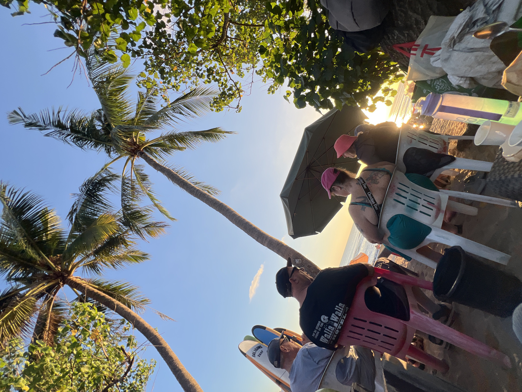 In Kuta, Indonesia, a group of individuals are seated on plastic chairs on a sandy beach, facing the ocean. The setting sun casts a warm glow on the scene, with palm trees silhouetted against the sky. Two surfboards lean against a railing in the background.
In the foreground, four people are visible. Two men in baseball caps are seated, one with a black t-shirt that reads "Walla Walla Hotel." To their right, two women in hats are also seated. One woman wears a teal bikini top and a black strap with "Christian Dior" visible.
An umbrella is open above the seated individuals, providing shade. To the right, bags and a water bottle are placed on the sand. The ocean is visible in the distance, with faint silhouettes of people on the beach. The image captures a relaxed moment as the day ends on the beach.