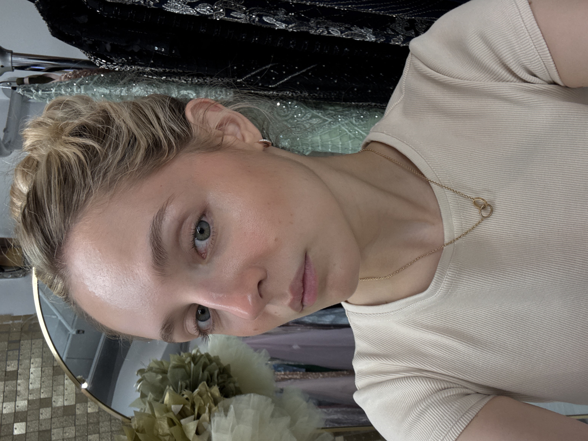 A young Caucasian woman with blonde hair pulled up in a bun, wearing a cream-colored ribbed t-shirt and a gold chain necklace with interlocking rings, is pictured in a selfie. She has on subtle makeup, with rosy cheeks and mascara. Her eyes are green and her expression is neutral. She is wearing small hoop earrings. In the background, to her left, is a round mirror with a gold frame, with a decorative display of gold and white fabric flowers. To her right, hanging on a rack, are several sequined dresses and other garments. The scene appears to be an indoor setting, possibly a boutique or a dressing room. The image does not provide specific information about the location within New York City.