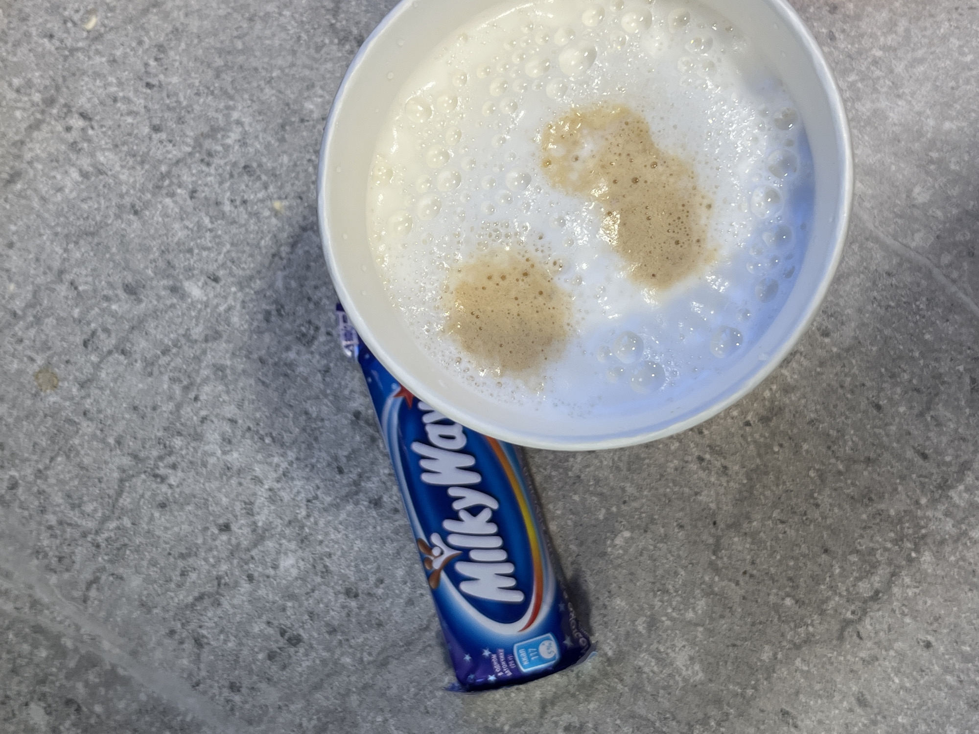 An overhead shot displays a white bowl or mug containing a frothy beverage, likely a latte or cappuccino, positioned on a mottled grey surface. The drink features a thick layer of white foam with numerous small bubbles, and two distinct, irregularly shaped patches of lighter brown liquid visible through the foam. Partially obscured beneath the bowl, and oriented diagonally, is a "Milky Way" chocolate bar in its blue wrapper. The wrapper prominently features the brand name "MilkyWay" in white and blue lettering, alongside a small brown illustrative logo. Additional text in Cyrillic script is visible, along with nutritional information indicating "117 kcal" and "6%". The underlying surface is a textured, grey material, resembling a stone or concrete countertop. No people are present, suggesting an intimate, indoor setting, possibly a kitchen or cafe. The presence of Cyrillic text is consistent with the provided location context of Oktyabrsky, Russia.