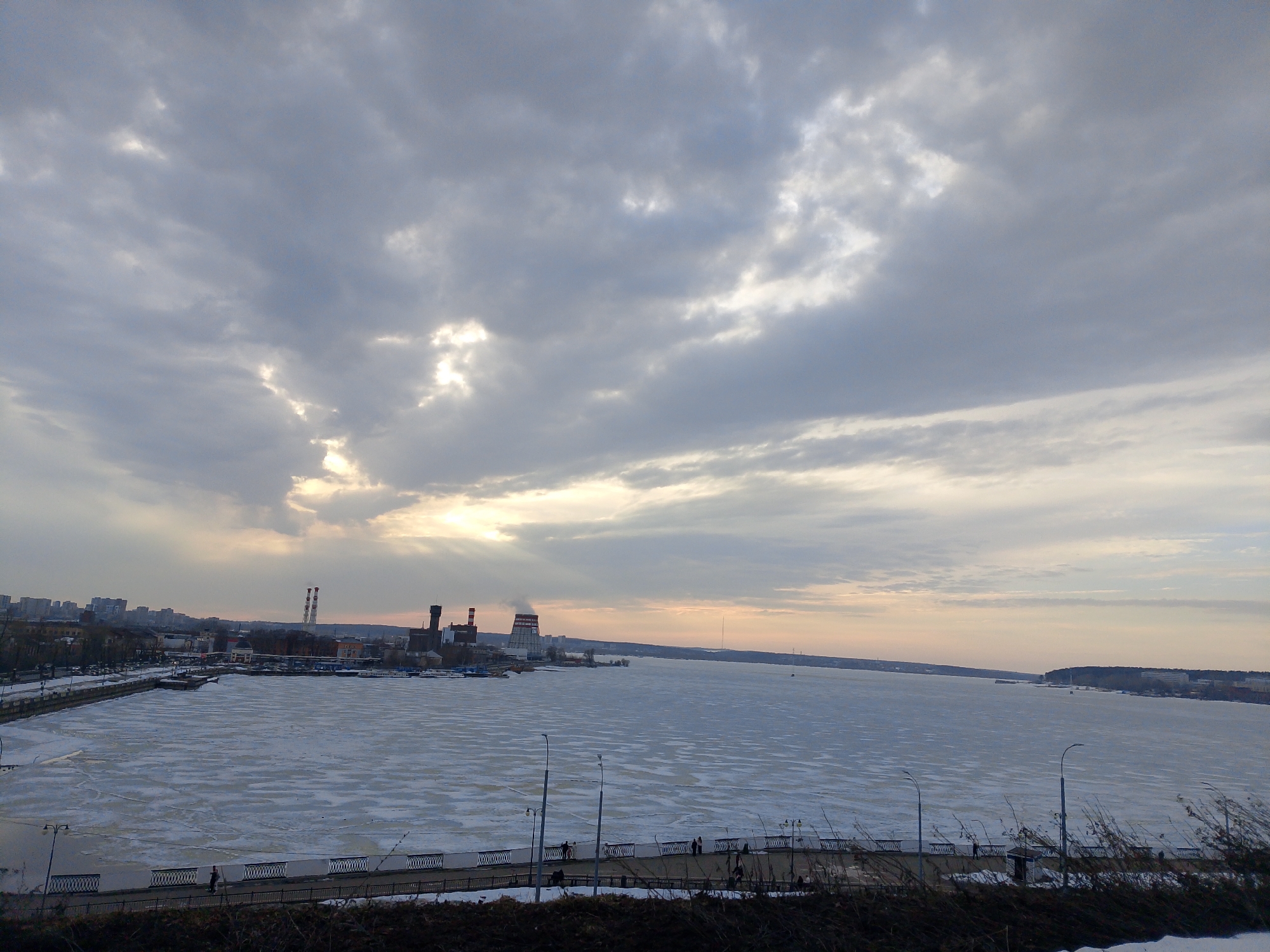 The image depicts a winter scene in Izhevsk, Russia, overlooking the frozen Izhevsk Pond. The sky is overcast with gray and white clouds, with a hint of sunlight breaking through. In the background, the city skyline is visible, including industrial structures such as chimneys and buildings. The Izhevsk Pond is covered with ice and patches of snow. A promenade runs along the edge of the pond, with streetlights lining the path. Several people are walking on the promenade. In the foreground, there is some vegetation.