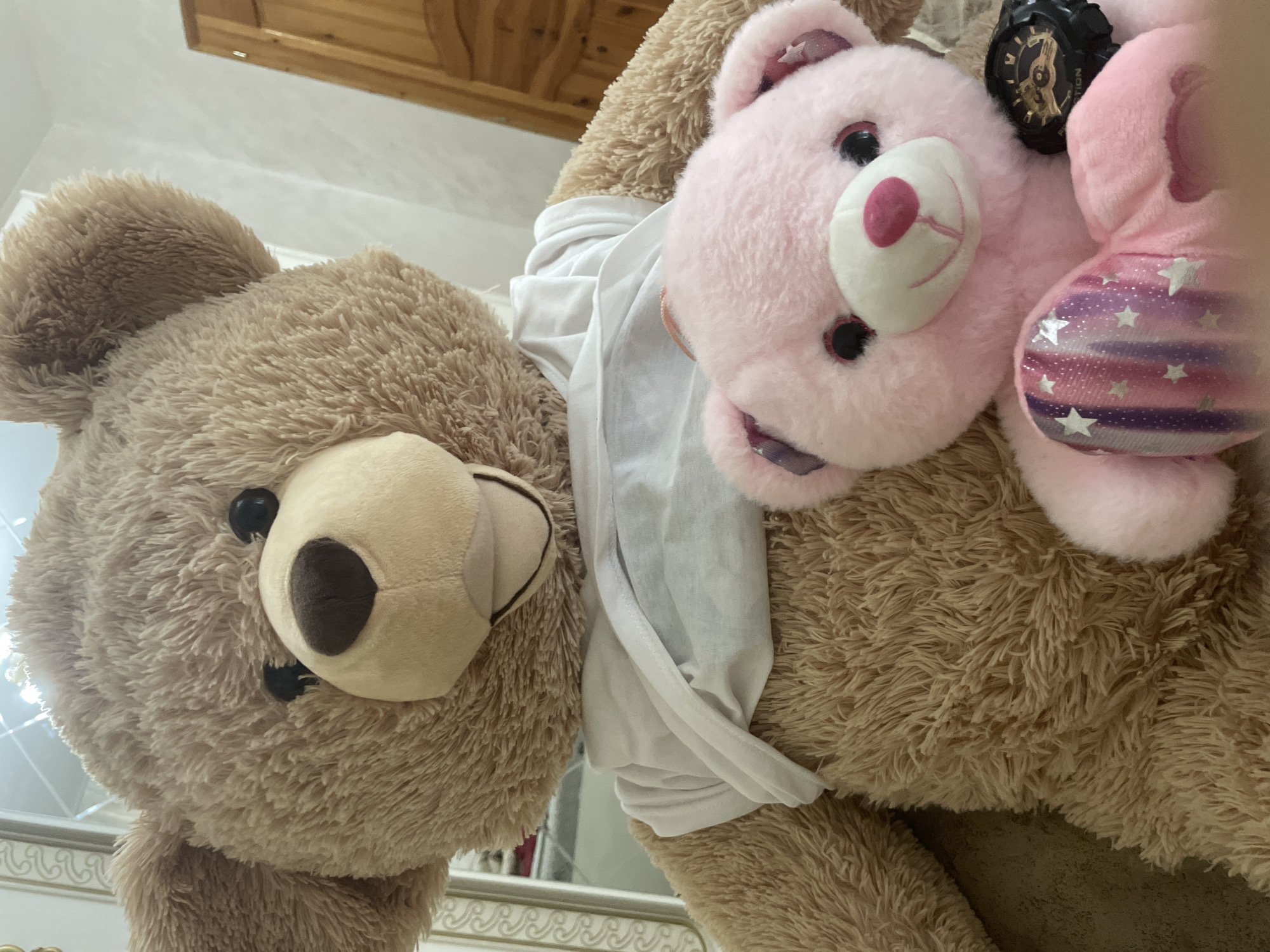The image captures two plush teddy bears in what appears to be an indoor setting, likely a bedroom, in Ihtiman, Bulgaria. The photo is taken from a low angle, looking upwards at the subjects.
In the foreground, a large, shaggy brown teddy bear dominates the left side of the frame, facing slightly towards the viewer with dark button eyes and a light brown snout. It appears to be wearing a plain white fabric, possibly a shirt, draped around its body. Nestled beside the large brown bear on the right is a smaller, bright pink teddy bear, lying on its back. This pink bear has large black eyes and a white snout with a pink nose.
A black wristwatch with a dark face and gold-toned accents is positioned on or near the pink bear's head. Visible text on the watch face reads "TION". Further to the right, a segment of another small pink plush item is visible, adorned with white stars over a pattern of purple and pink stripes.
The background consists of light-colored, possibly white, walls and a wooden ceiling or beam structure at the very top of the frame. Below the bears, a white, ornately carved or decorative furniture piece, potentially a headboard or railing, is partially visible. The lighting is soft and ambient, characteristic of an indoor environment.