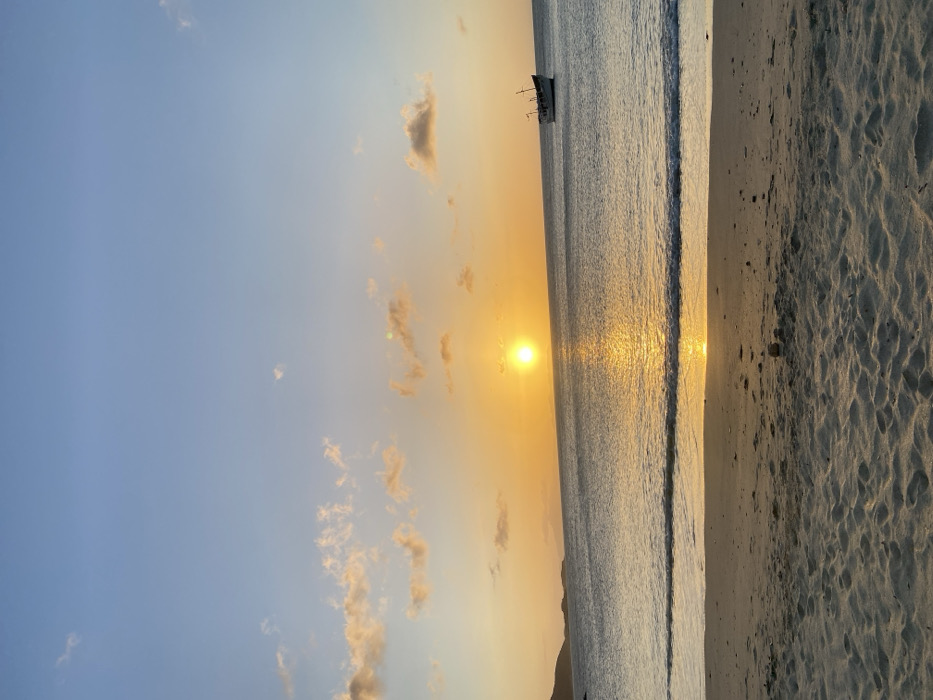This image depicts a coastal sunset scene in Juan Griego, Venezuela. The sky transitions from a light blue at the top to a vibrant orange-yellow hue near the horizon, where the sun is visible as a bright, yellow-orange orb slightly left of center. Scattered, illuminated clouds are present in the sky. The mid-ground features a calm body of water reflecting the sun's light, creating a bright path extending towards the viewer. A small, light-colored vessel, possibly a fishing boat or sailboat with visible rigging, is positioned on the water in the right portion of the frame. Distant landmasses are silhouetted on the left horizon. The foreground consists of a sandy beach with subtle textures and gentle waves breaking at the shoreline. No individuals or specific actions are discernible.