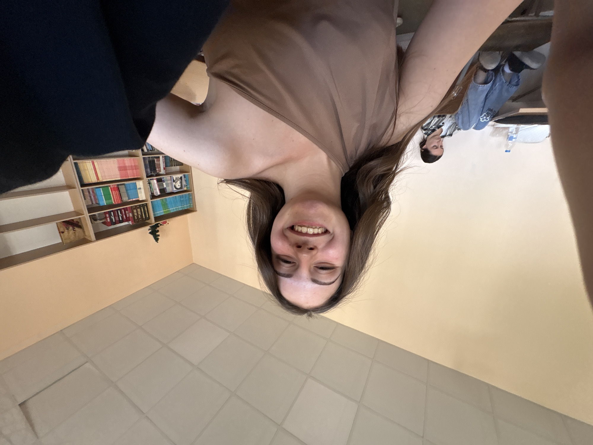The image is an upside-down selfie of a smiling young woman with brown hair and braces. She wears a brown sleeveless top. Behind her is a bookshelf filled with books, a tiled floor, and a peach-colored wall. In the background, a person wearing jeans and a black-and-white striped shirt is sitting at a table with a water bottle on it. The location cannot be determined to be in Burgas, Bulgaria, from this image alone.