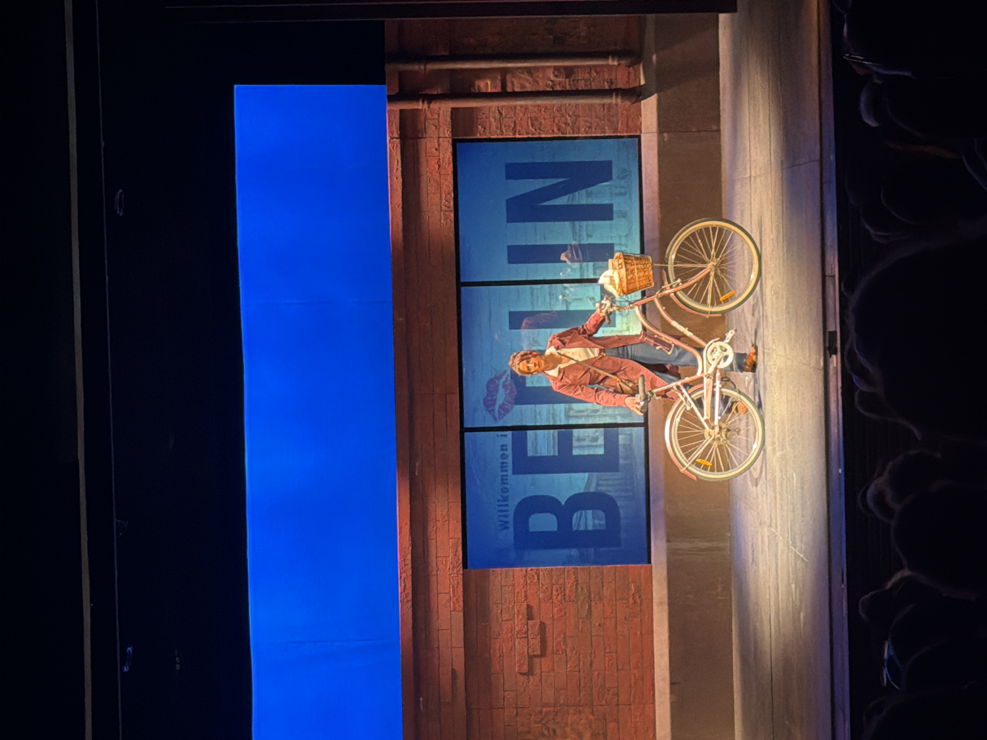 The image shows a stage production, likely a musical or play. The set depicts a brick wall with the word "BERLIN" displayed vertically in large blue letters. Below the word, "Willkommen in" is visible. A person dressed in a red suit is riding a bicycle with a basket on the front. The scene is set against a blue backdrop. The location of the image is not the city of Tel-Aviv, Israel.
