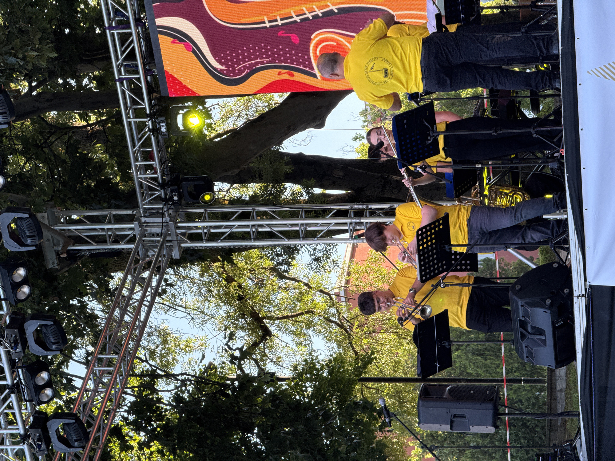 An outdoor daytime event is underway on a stage. Multiple individuals, mostly wearing bright yellow t-shirts, are performing music. Two young men on the left are playing trumpets, and a young woman in the center is playing a flute. A man in a yellow t-shirt is facing away from the camera, looking towards the musicians. A large screen with a colorful, abstract design is visible on the right side of the stage. Stage lighting trusses and speakers are also present. The background features trees and what appears to be a building. The scene suggests a musical performance or festival. The logo on the back of the yellow shirts is visible and contains text that is likely related to the city of Pleven, Bulgaria, possibly indicating this event is taking place there.